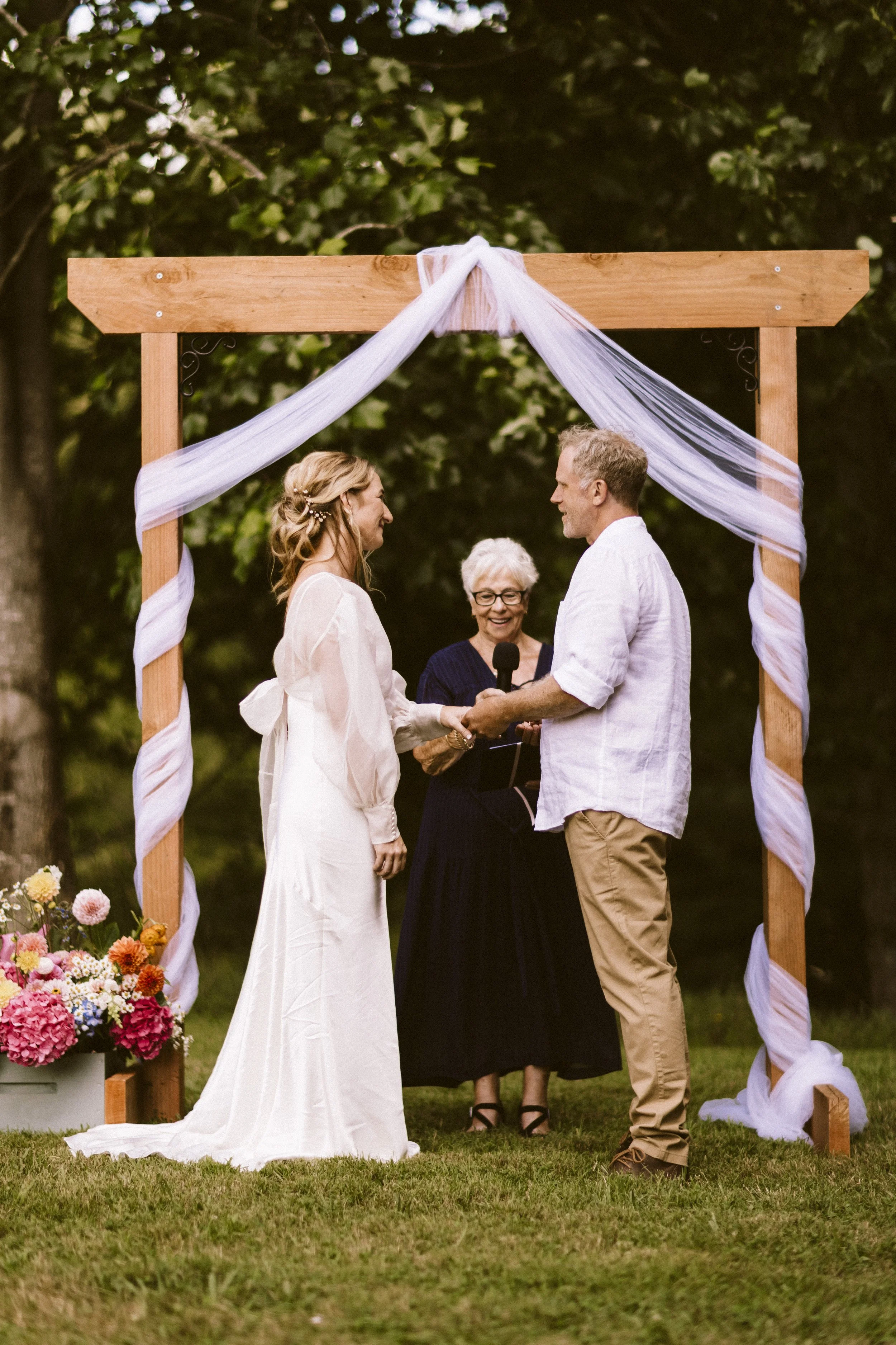 A couple exchanging vows at an outdoor wedding ceremony under a wooden arch decorated with white fabric, with a officiant standing behind them and a basket of colorful flowers nearby.