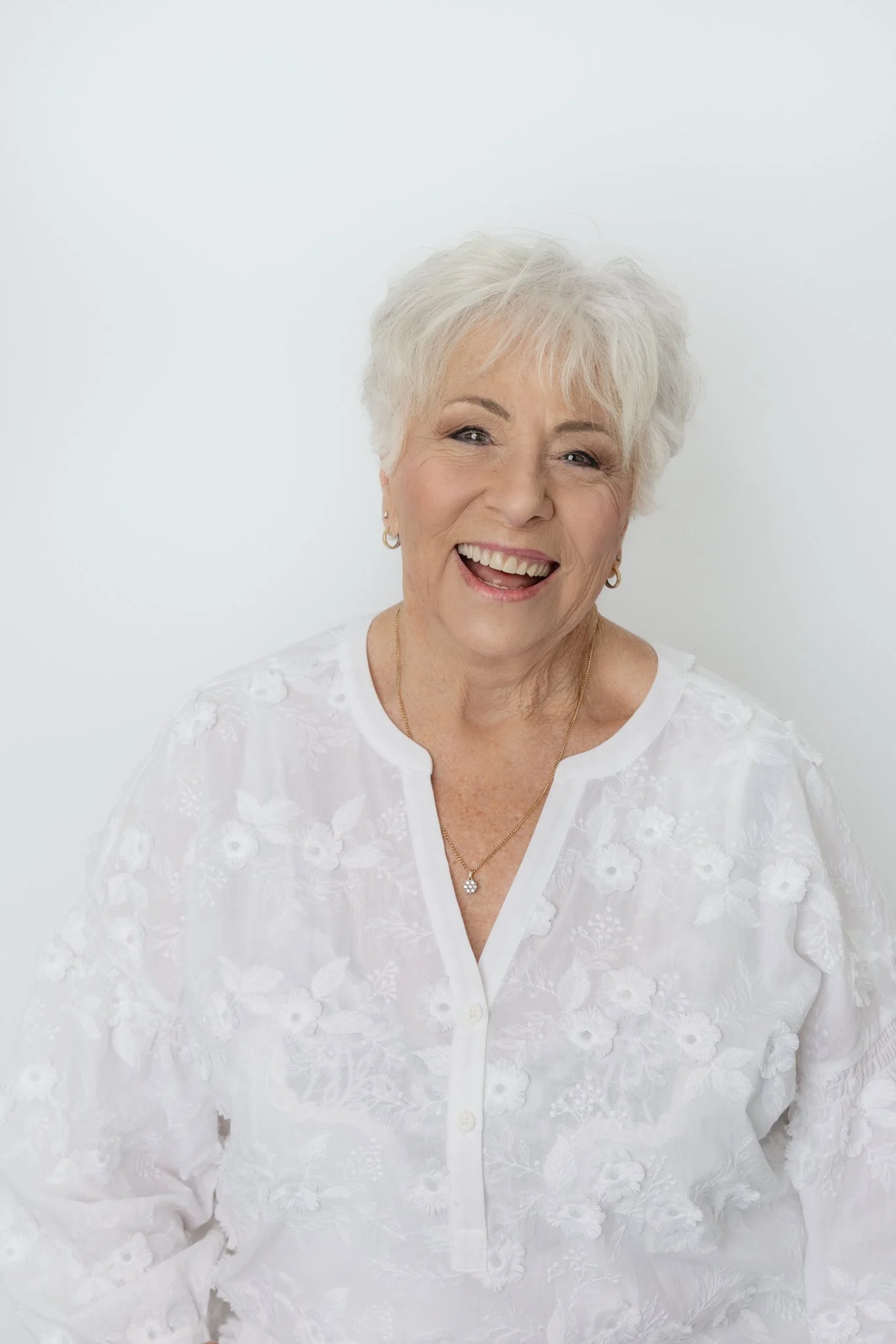 Smiling elderly woman with short white hair wearing a white embroidered blouse, gold earrings, and a gold necklace, against a white background.