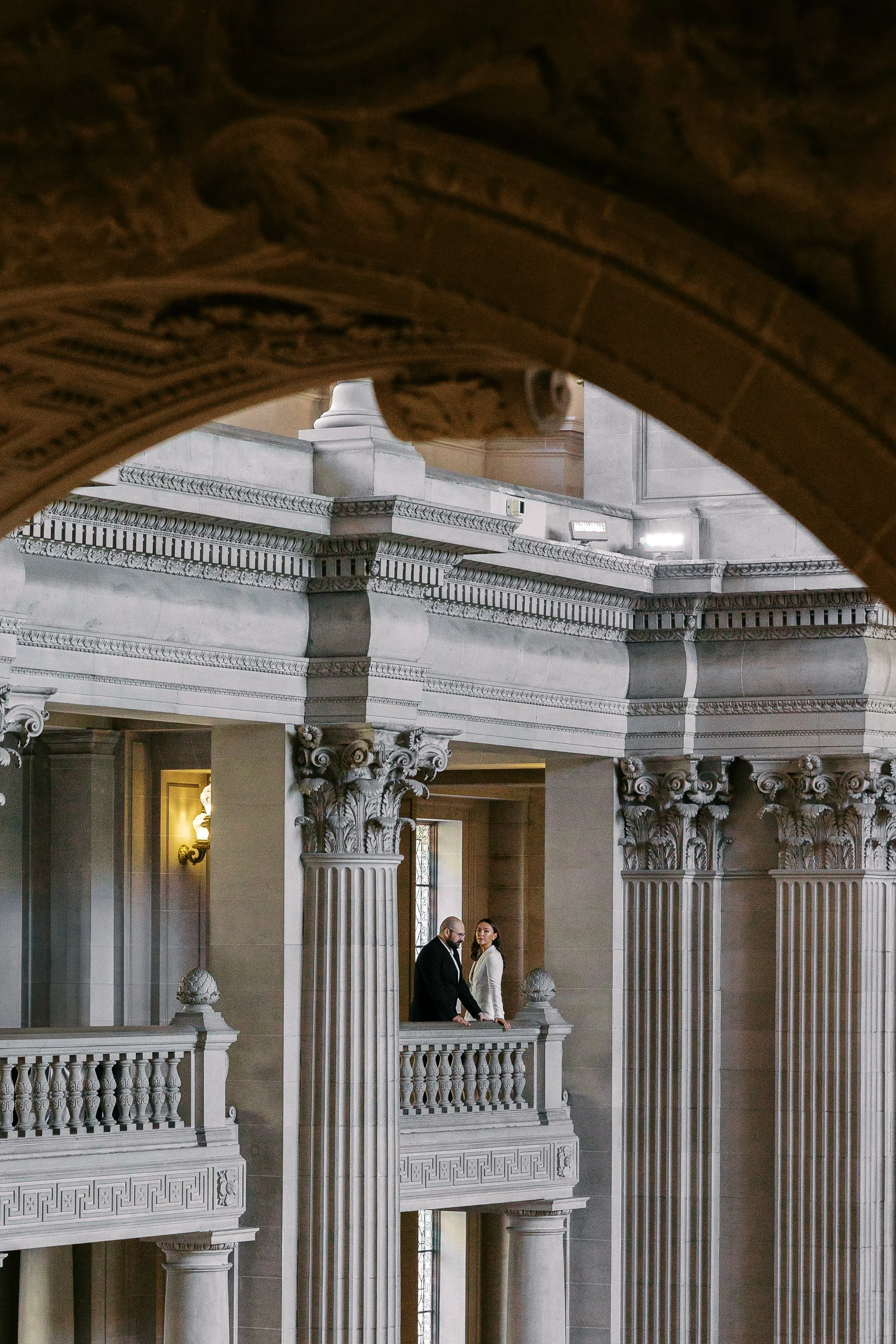 Two people standing on a balcony in an ornate, classical building with columns and detailed architectural elements.