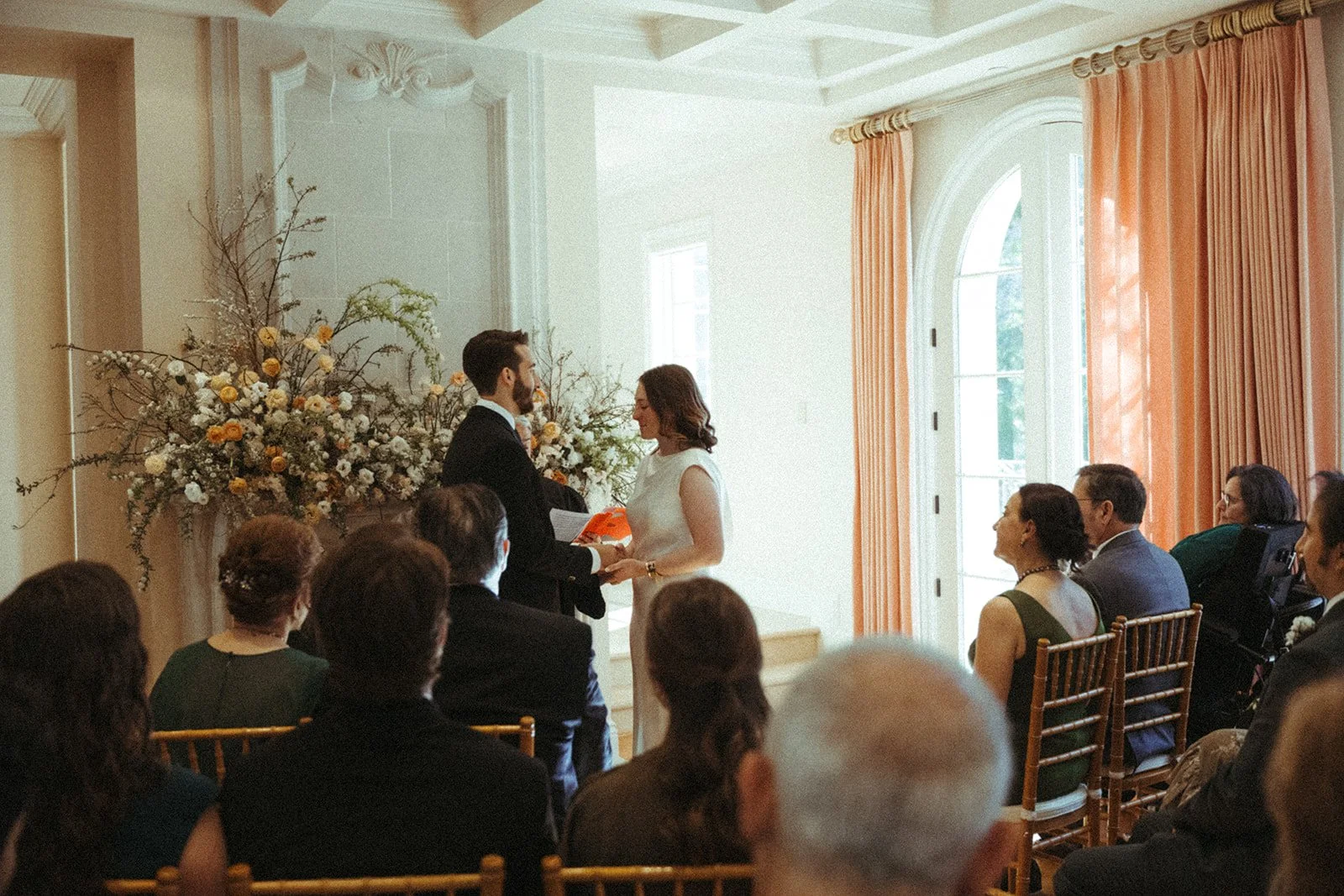 A wedding ceremony with a bride and groom standing together, facing each other, holding hands. The bride is wearing a white dress, and the groom is in a black suit. They are in a decorated room with peach curtains and large windows letting in natural light. Guests are seated in chairs, watching the ceremony, with a large floral arrangement behind the couple.