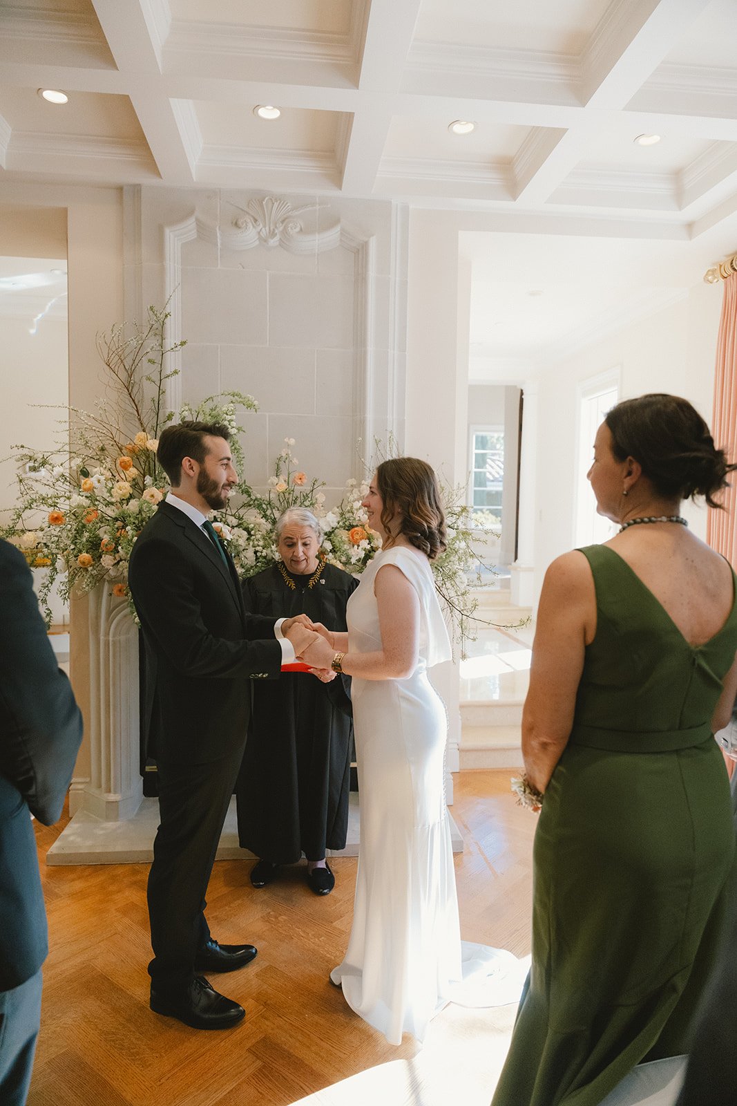 A wedding ceremony with a bride and groom exchanging vows inside a bright, elegant room with floral decorations, attended by a woman in a green dress.