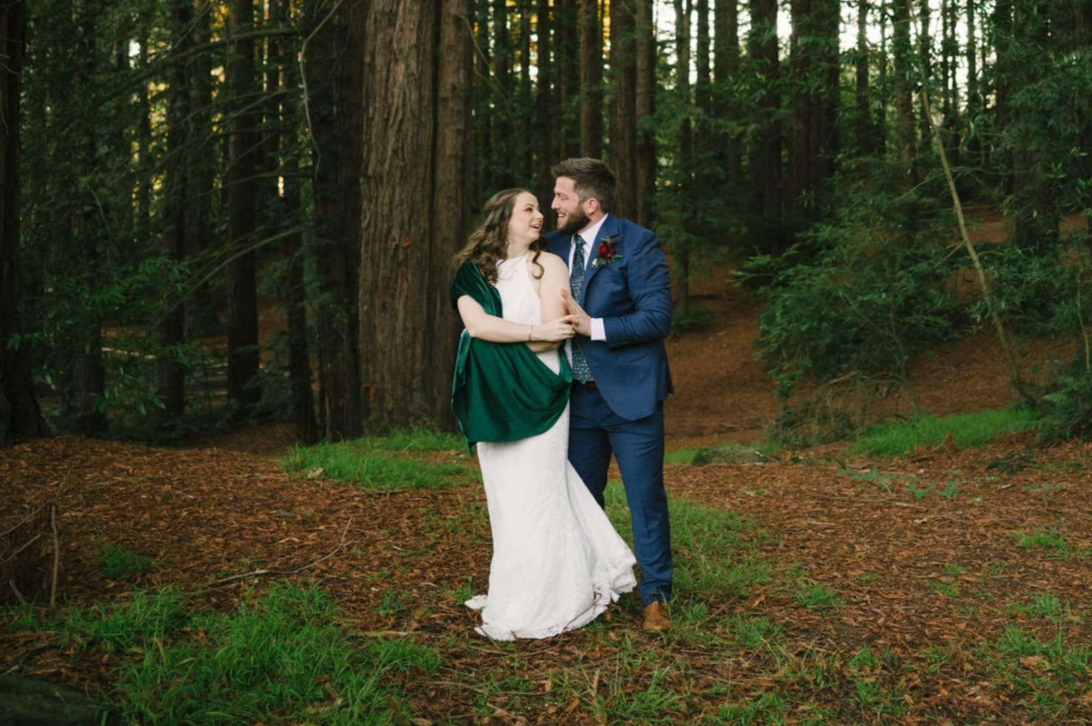 A couple dressed in wedding attire standing close together in a forest with large trees and green foliage, sharing a joyful moment.