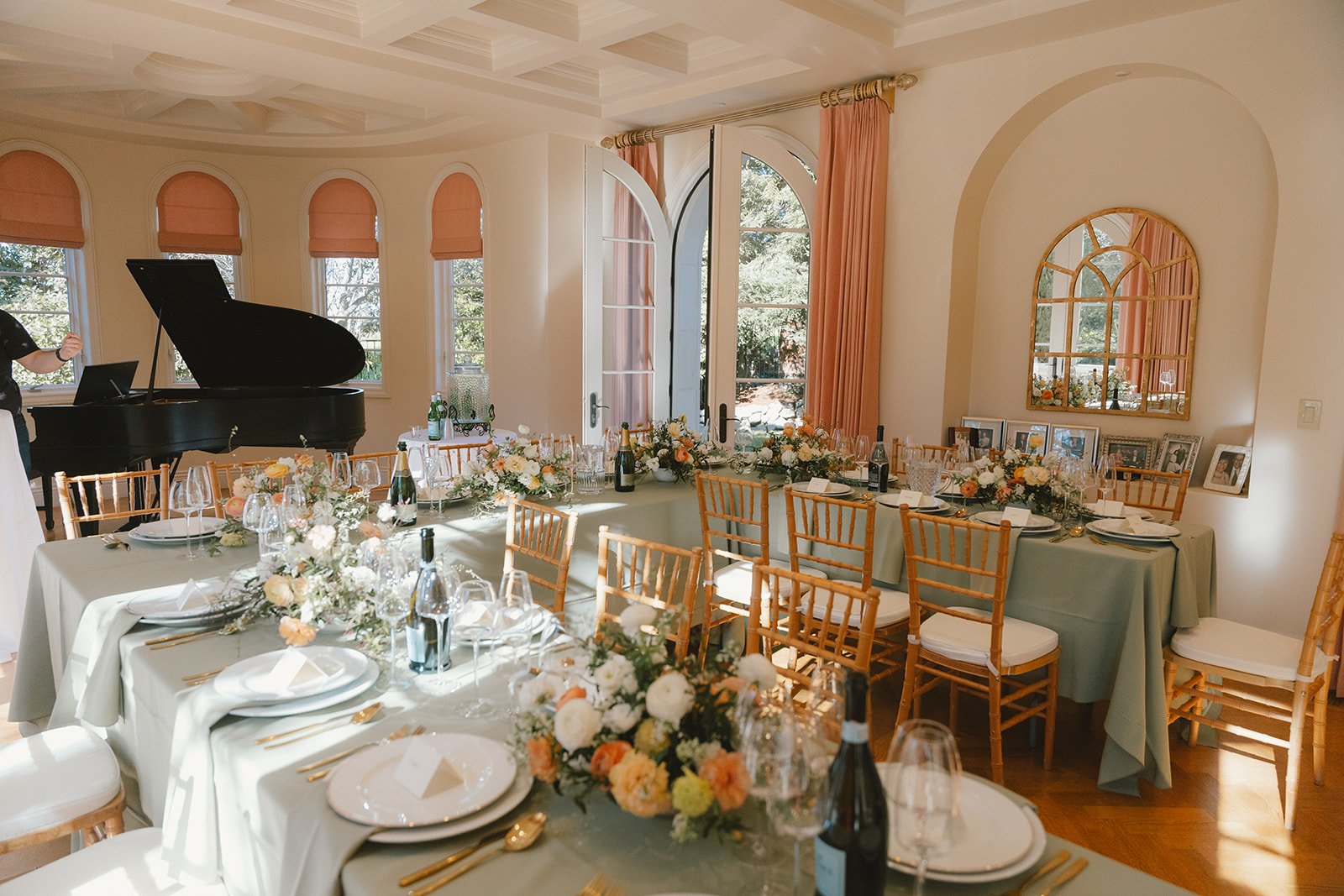 Elegant dining room with rectangular tables decorated with floral centerpieces, surrounded by gold chairs, set for a meal, with a grand piano in the background and large arched windows and doors letting in natural light.