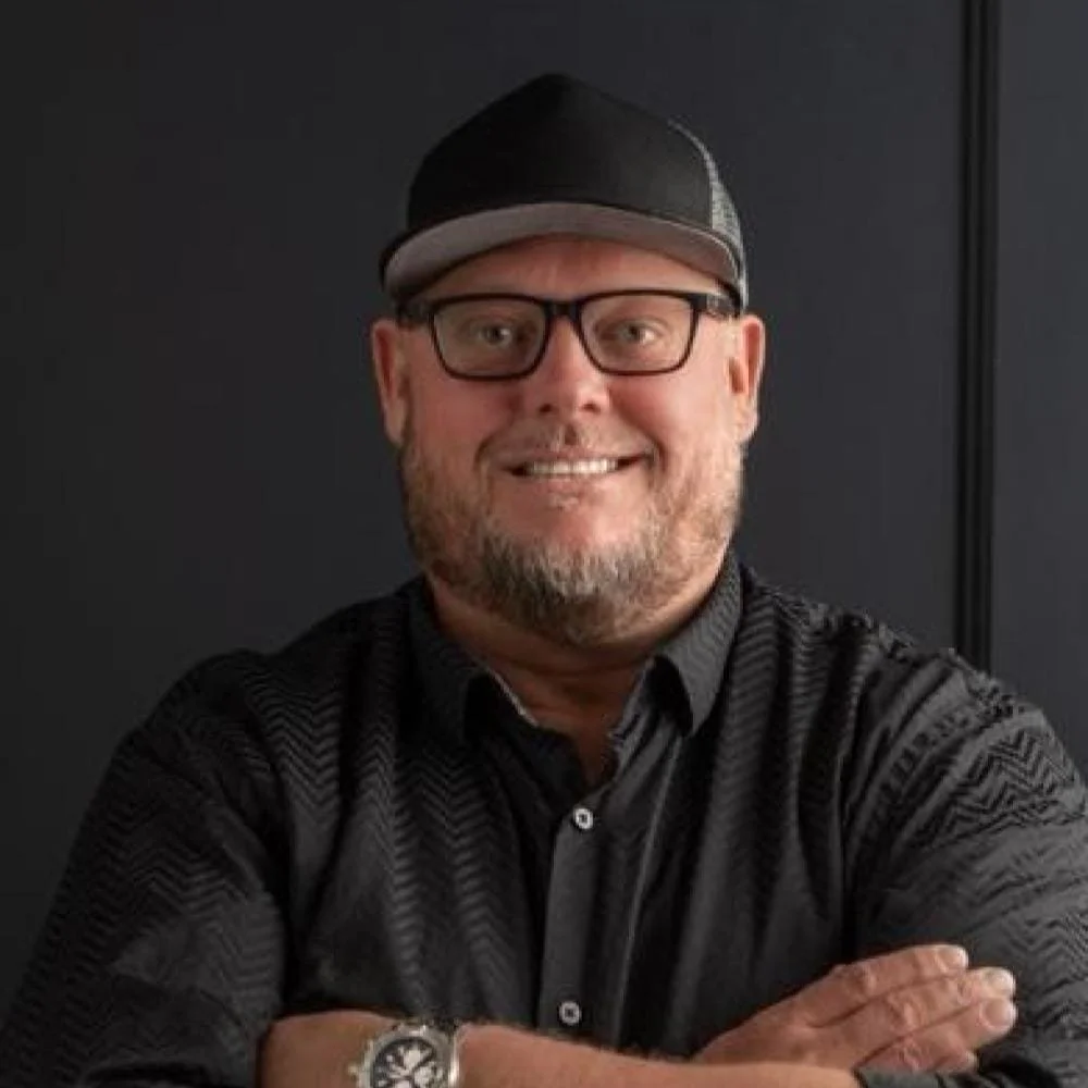 Photo of featured celebrity chef Beau MacMillan. Man with glasses and beard wearing a black baseball cap and black button-up shirt, smiling at the camera, with crossed arms against a dark background.