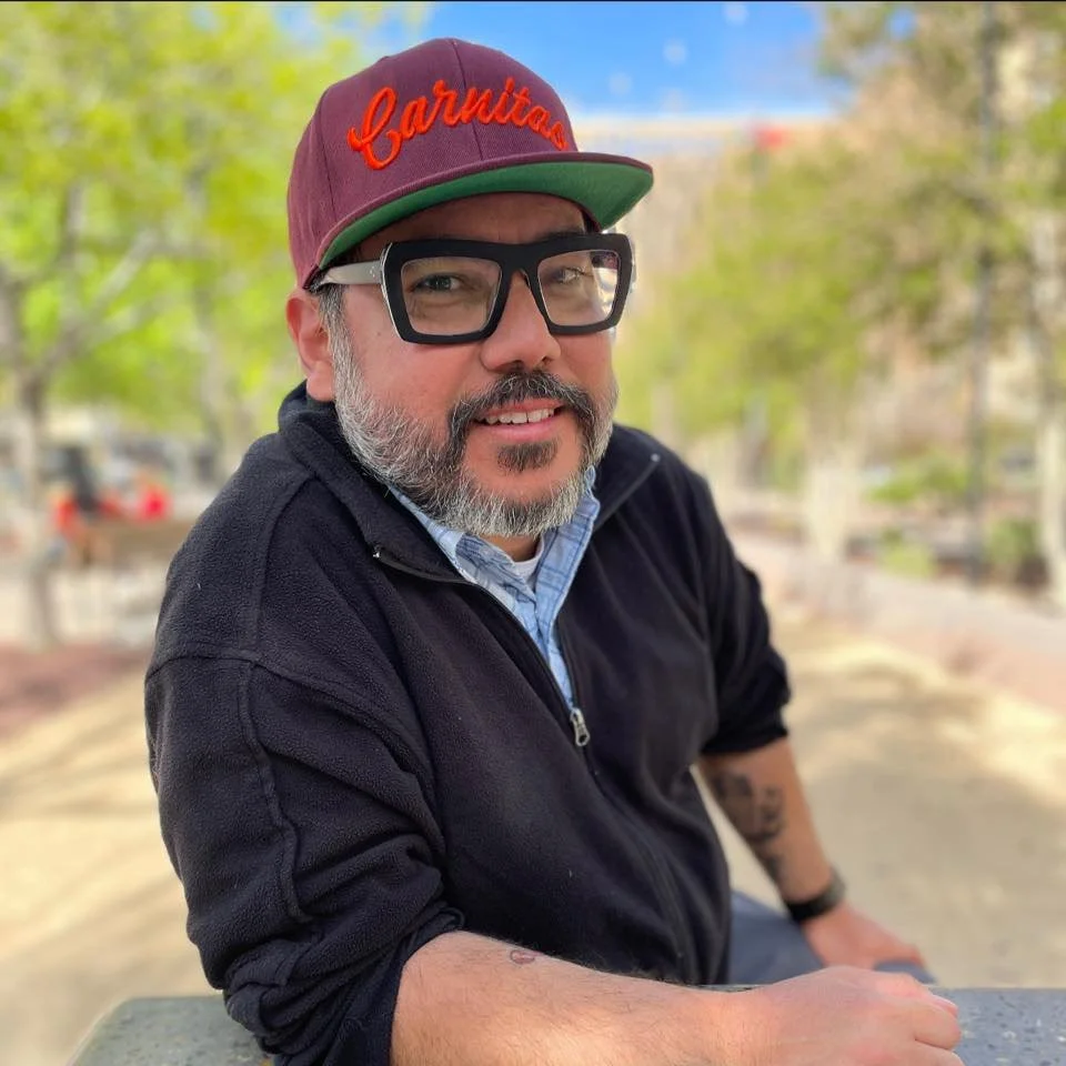 A man sitting outdoors at a park or outdoor area, wearing a maroon baseball cap with red embroidered text, large black glasses, a black zip-up jacket, and a blue collared shirt underneath. He has a beard and mustache, and is smiling at the camera. The background shows green trees and a blurred park setting.