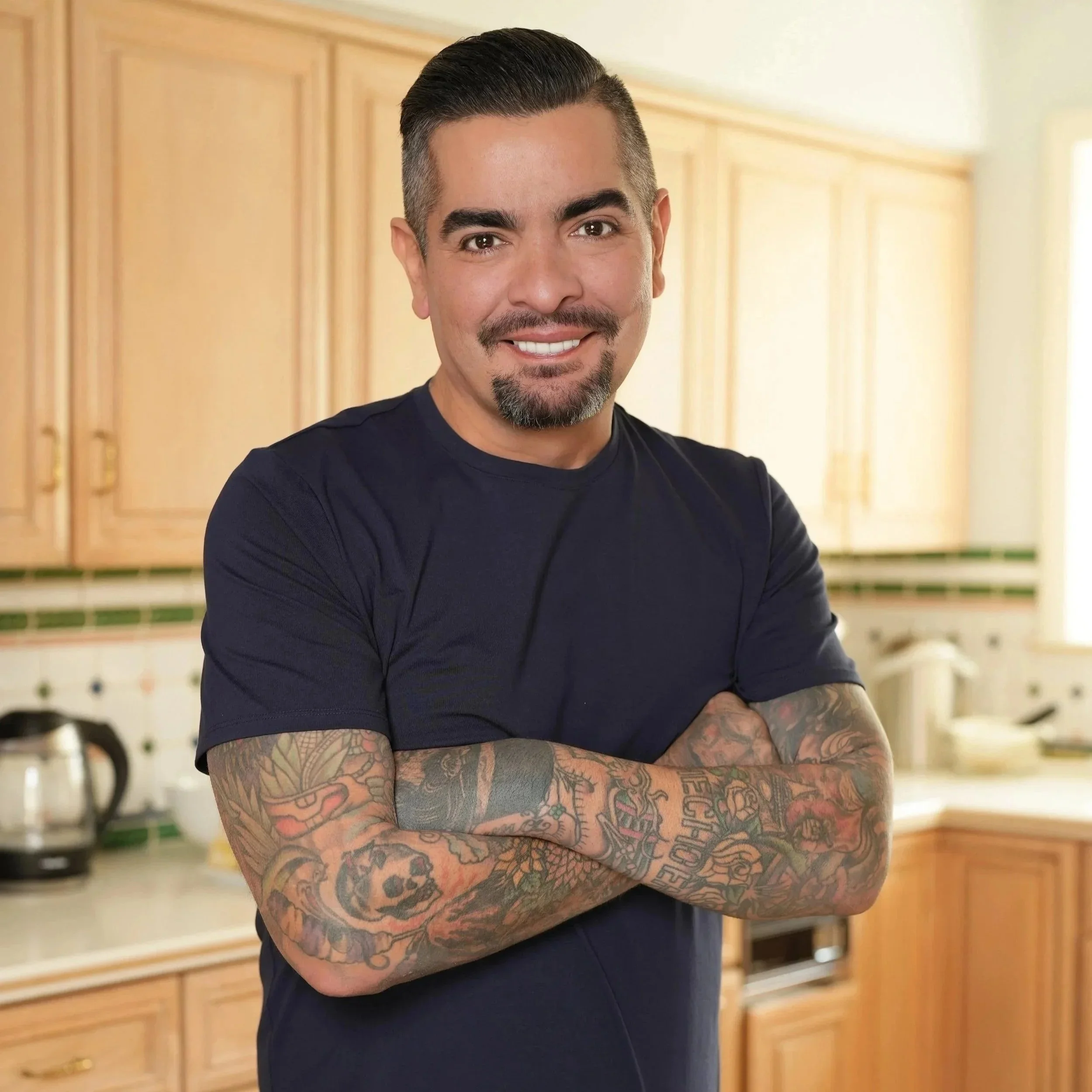 Photo of featured celebrity Chef Aarón Sánchez. A man with styled dark hair, a beard, and tattoos on his arms, smiling and crossing his arms in a kitchen with wooden cabinets.
