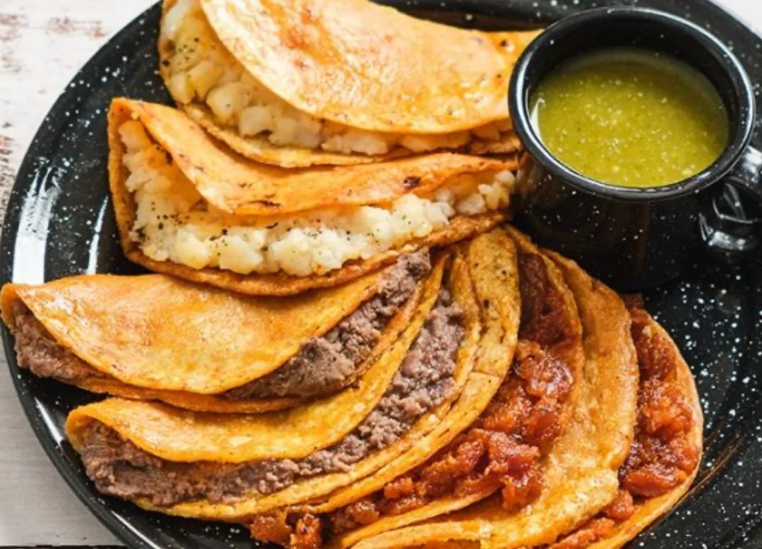 Plate of assorted Mexican tacos with beef, potato, and other fillings, and a small cup of green salsa.