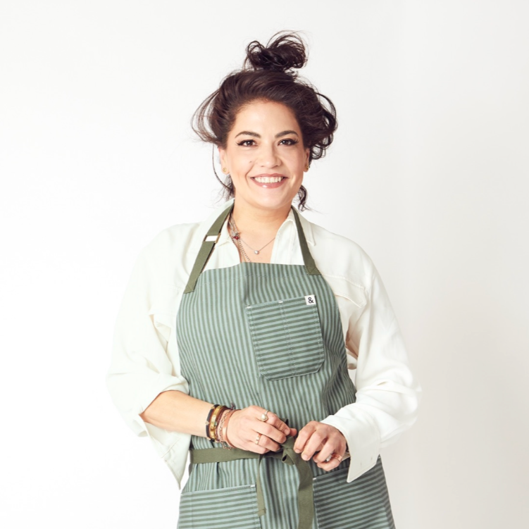 Featured Chef Maria Mazon of Boca Tacos in Tucson. Smiling woman with dark curly hair wearing a green striped apron over a white shirt standing against a plain white background.