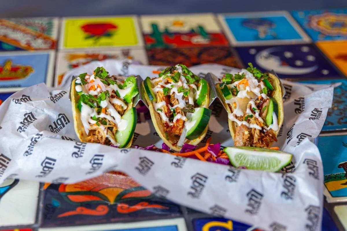 Three tacos filled with shredded meat, onions, cilantro, and drizzled with white sauce, garnished with cucumber slices and lime wedge, on branded paper, with colorful tiles in the background.