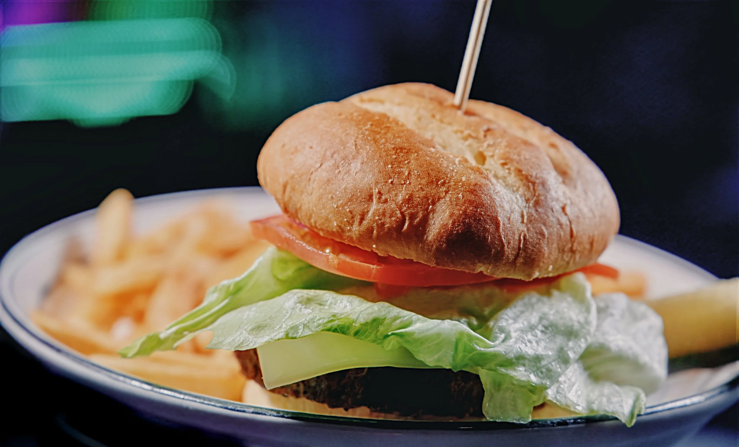 Close-up of a burger with lettuce, tomato, and a sandwich bun, served on a dark plate with French fries in the background.