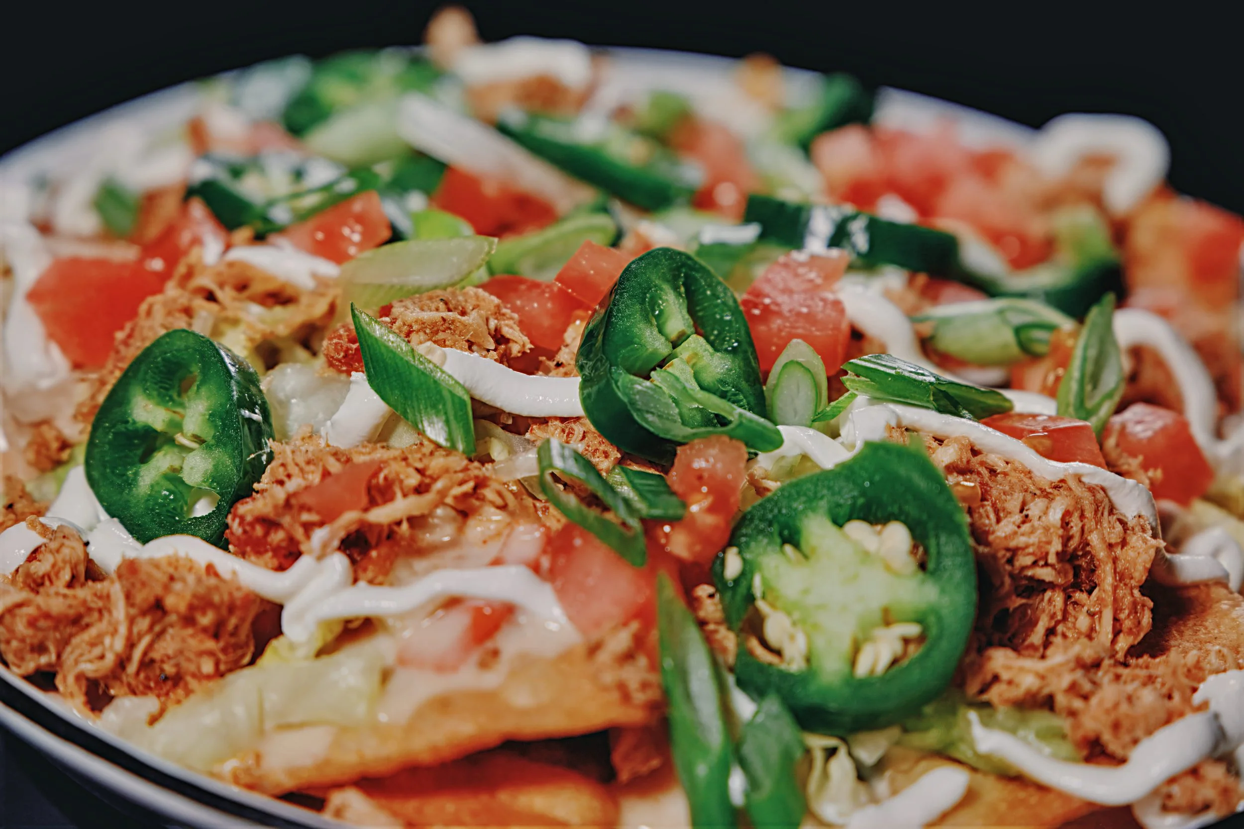 Close-up of a plate of nachos topped with shredded chicken, sliced jalapeños, chopped tomatoes, green onions, and drizzled with white sauce.