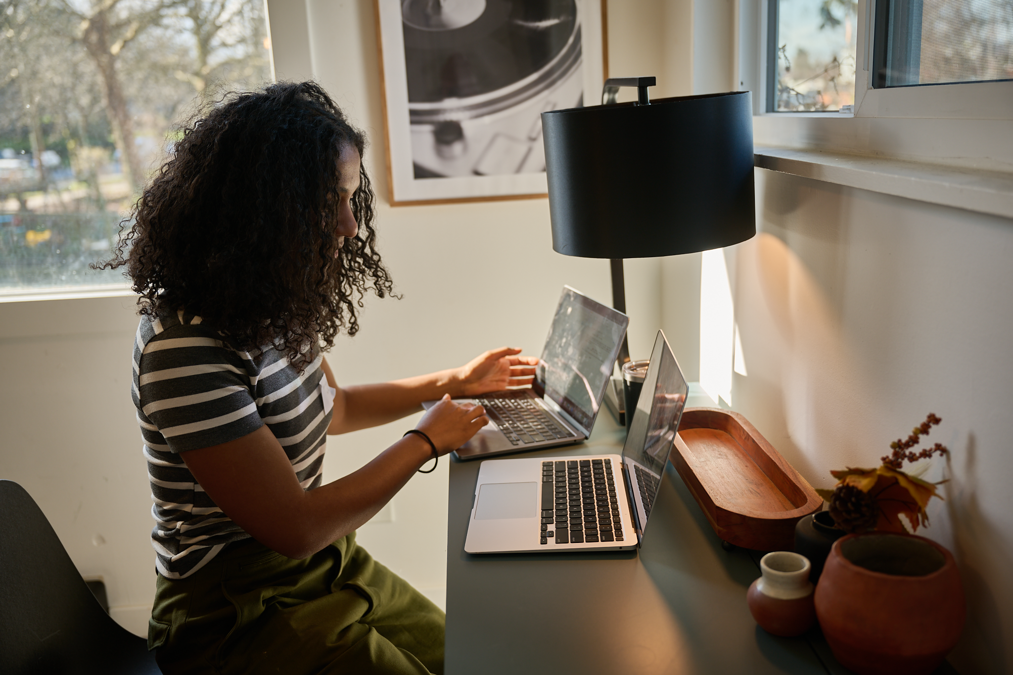 A woman with curly hair sitting at a desk working on two laptops, with a window to her right and decorative vases on the desk.