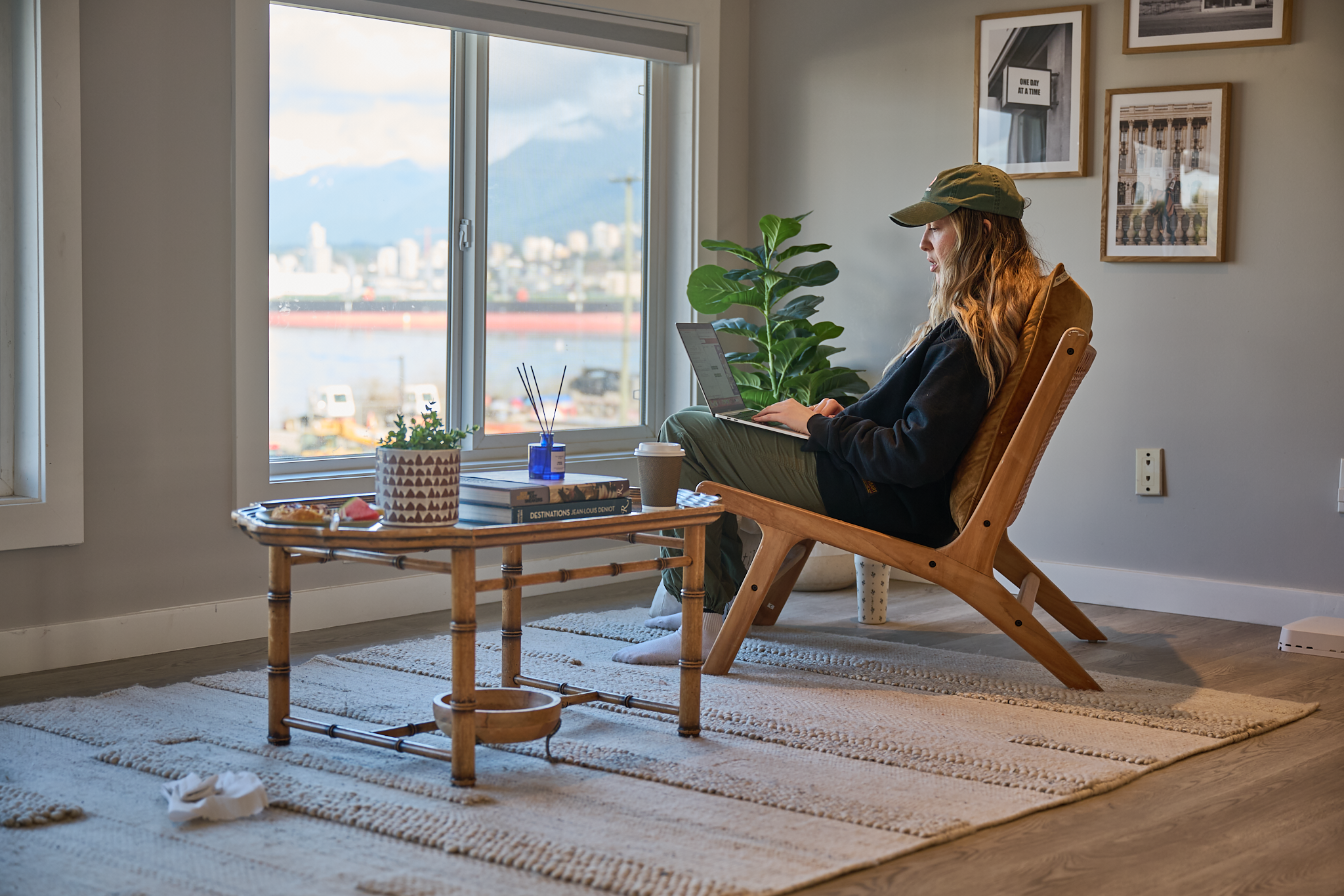 A woman with long wavy blonde hair wearing a green baseball cap, black jacket, and olive green pants sitting in a wooden lounge chair in a bright living room with large window overlooking city buildings and mountains in the distance. The room has framed black and white photographs and a large green plant next to her. On the coffee table are a plant in a patterned pot, books, a blue diffuser with sticks, a coffee cup, and a plate with snacks. The floor has a beige textured rug.