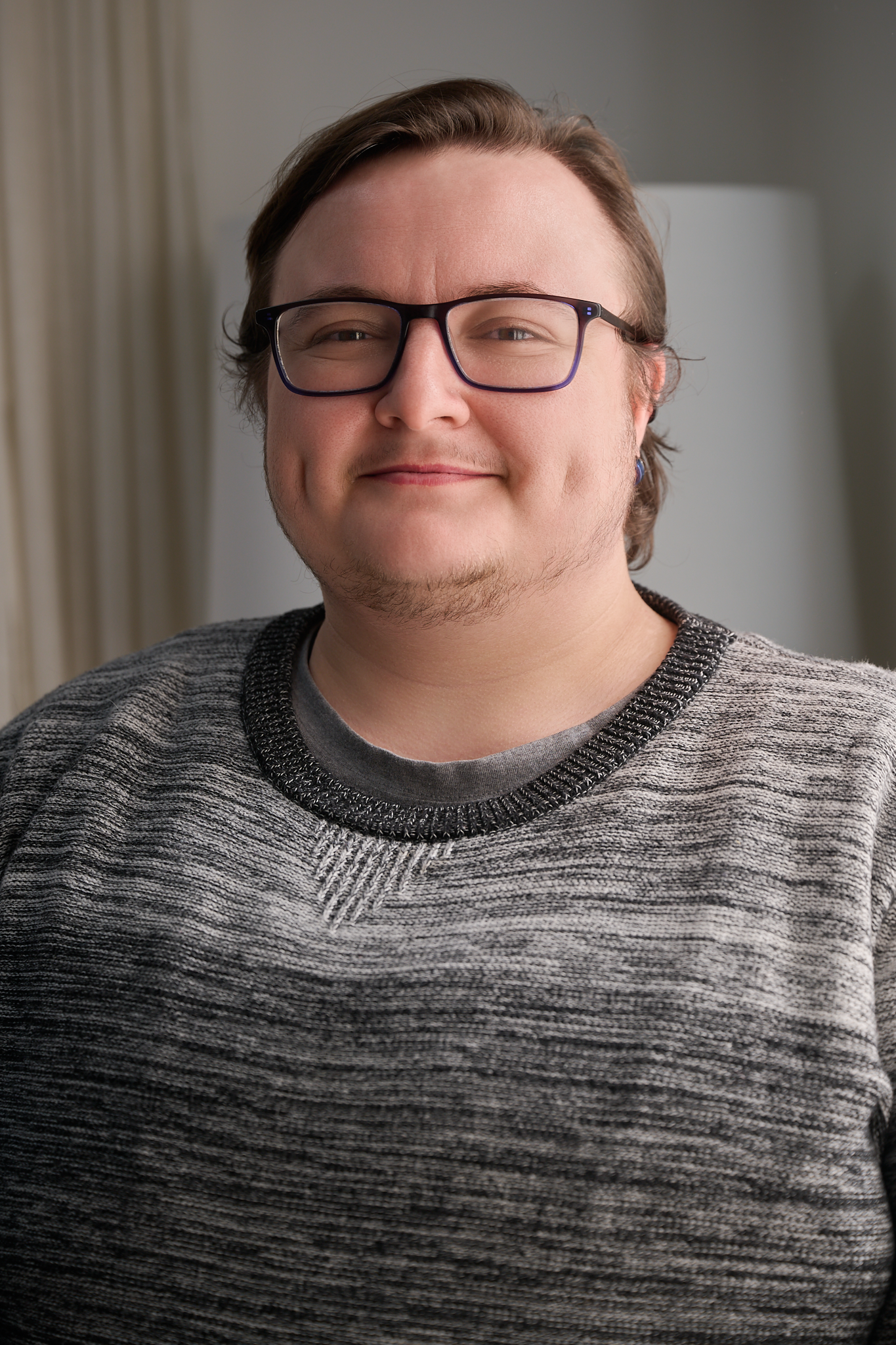 Portrait of a young man with glasses smiling, wearing a gray patterned sweater, indoors.