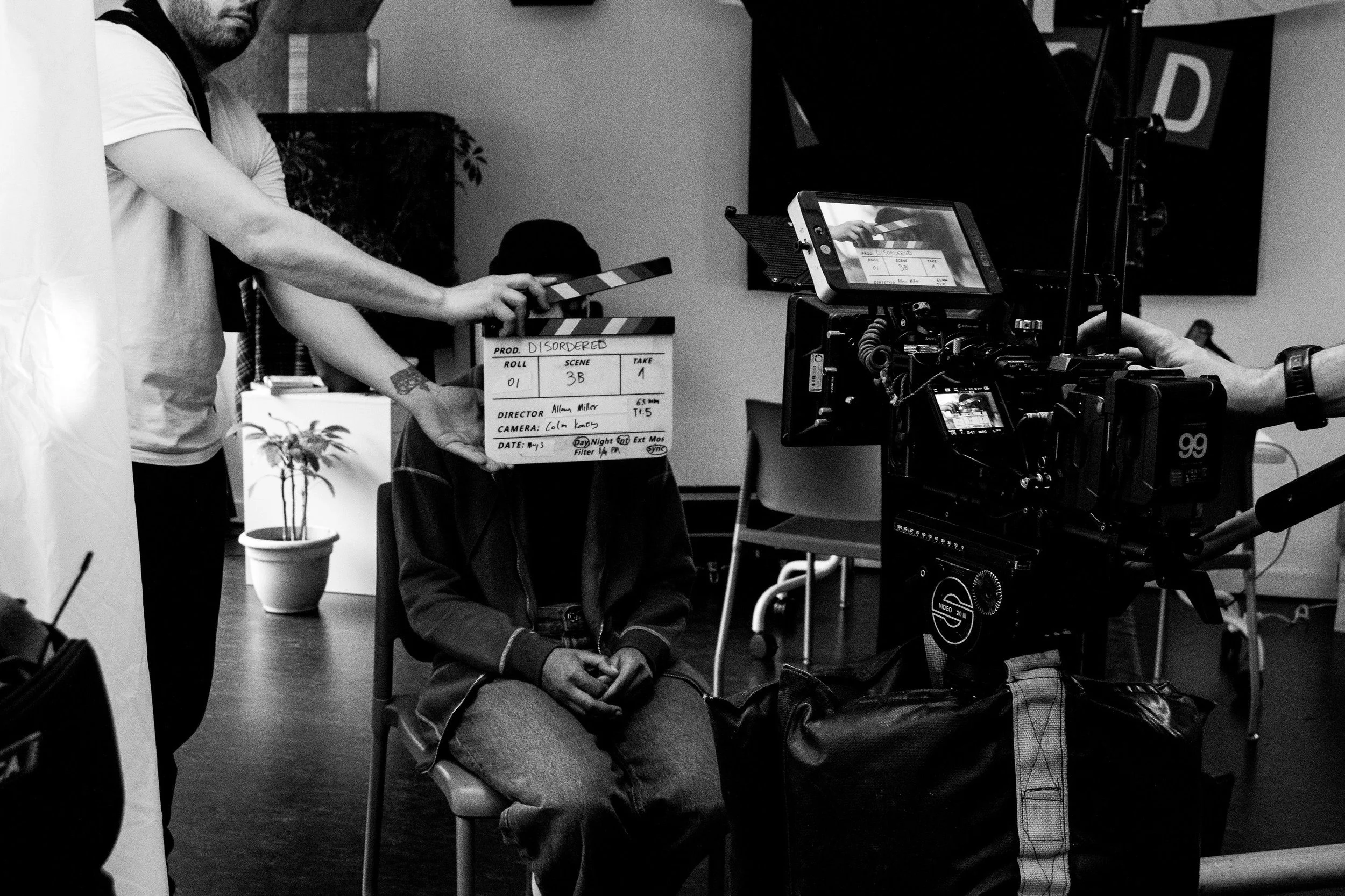 A black and white photo of a film production scene showing a person sitting in a chair with hands folded, a person standing to the left holding a clapperboard, and professional video equipment including a camera and monitor capturing the scene.