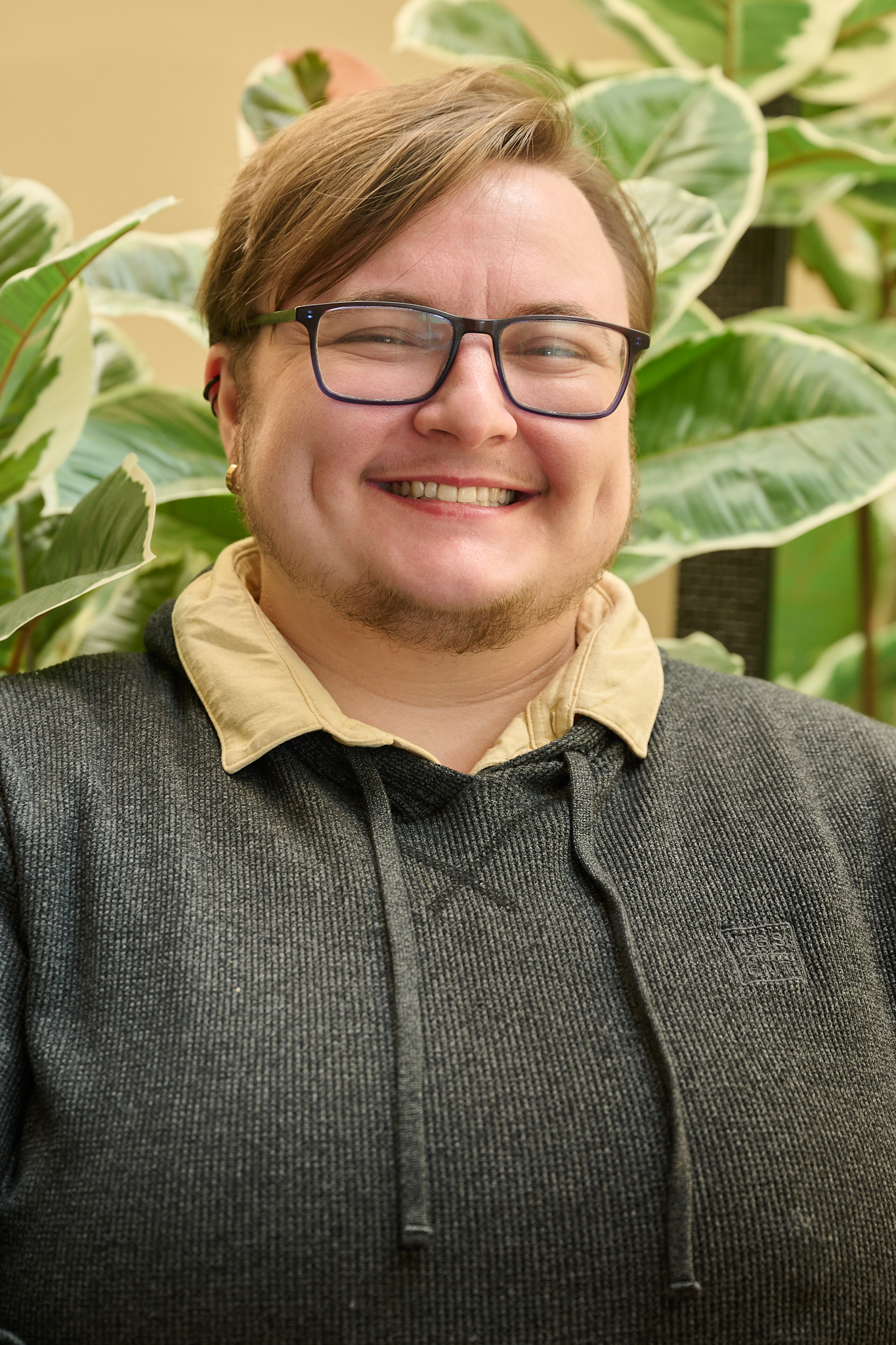 A smiling person with glasses, light brown hair, and a beard, wearing a hoodie and a collared shirt, standing in front of large green variegated leaves.