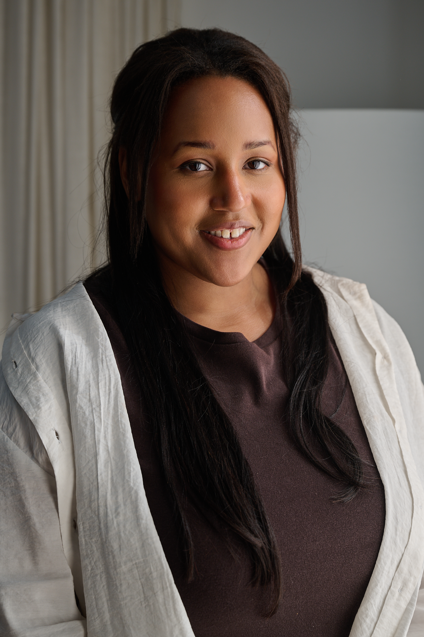 A young woman with long dark hair, wearing a brown shirt and a white jacket, smiling and looking at the camera in a well-lit indoor setting.