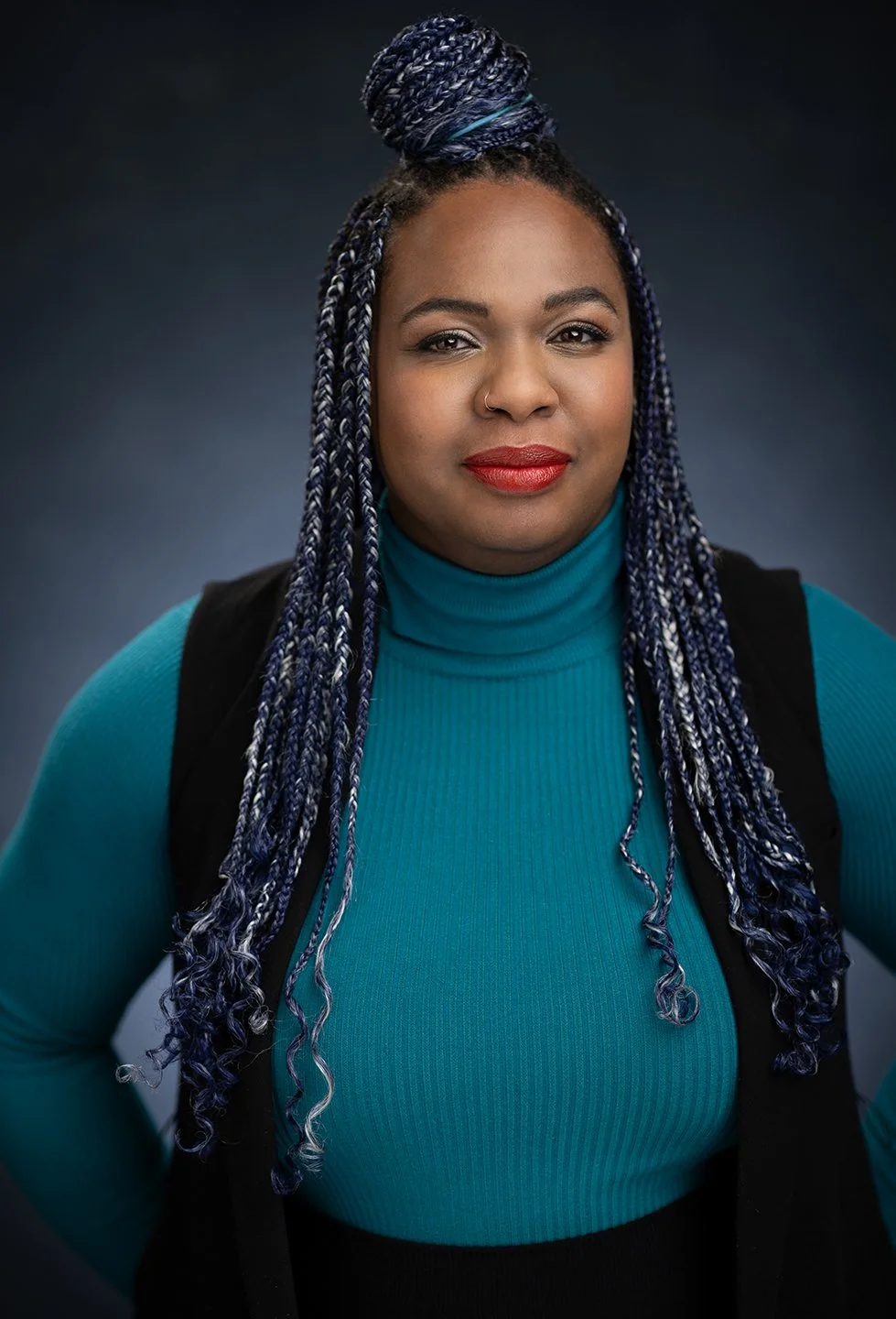 A woman with dark skin and long braided hair with blue and white highlights, wearing a teal turtleneck and a black vest, poses against a dark gradient background.