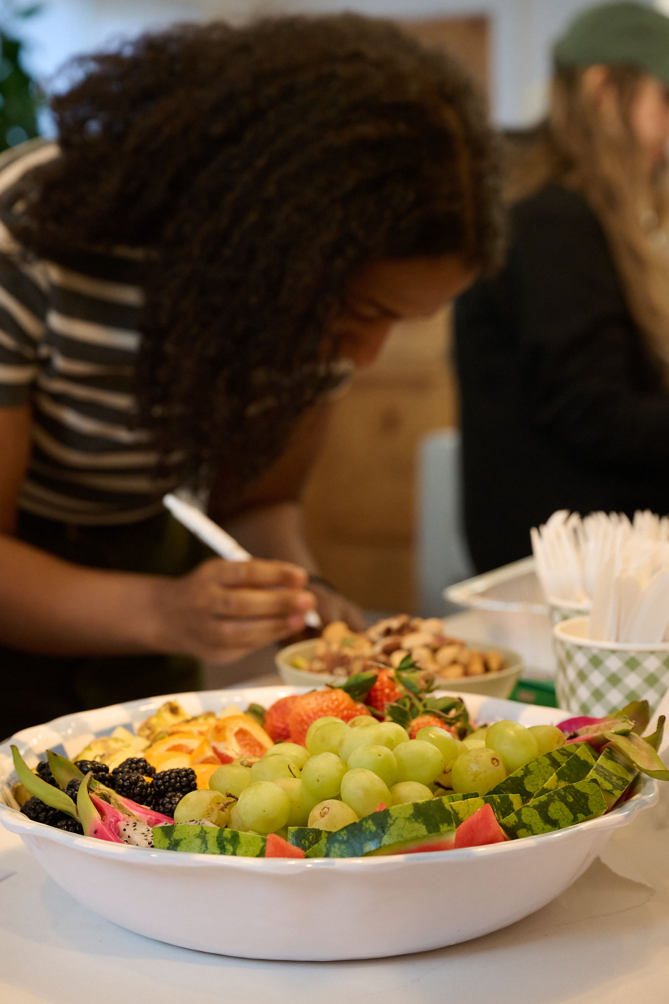 A woman with dark, curly hair and a striped shirt is looking down and writing, with a large bowl of assorted fruits including grapes and strawberries in the foreground.