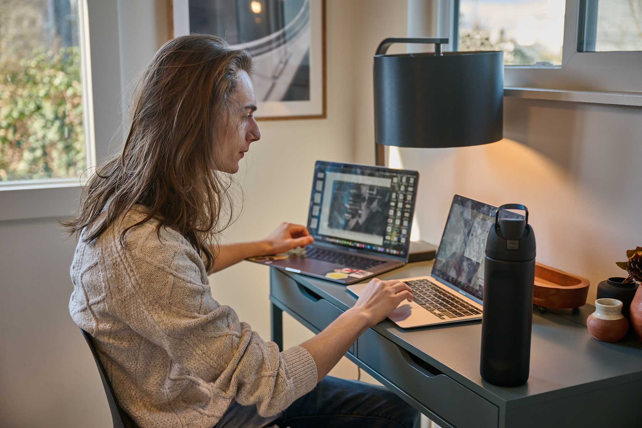 Man working at a desk with two laptops, a black water bottle, and decorative pottery in a well-lit room with large windows.