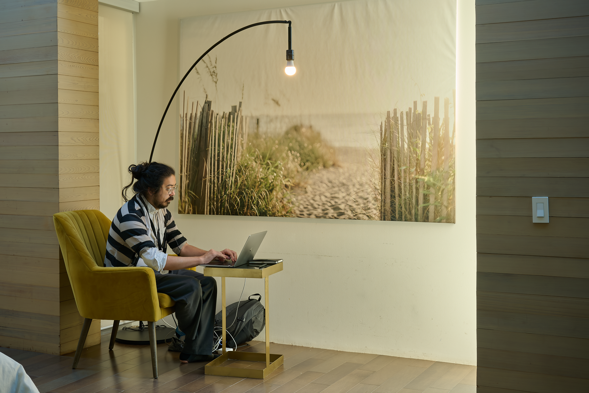 A man with dark hair and a beard, wearing glasses, a striped shirt, and dark pants, sitting on a yellow armchair and working on a silver laptop in a room with wood-paneled walls. Behind him, a large photograph of a sandy path leading through a beach with wooden fences and vegetation is hung on the wall. An arc floor lamp with an exposed bulb is positioned above the man, and a gray backpack is on the floor beside him.