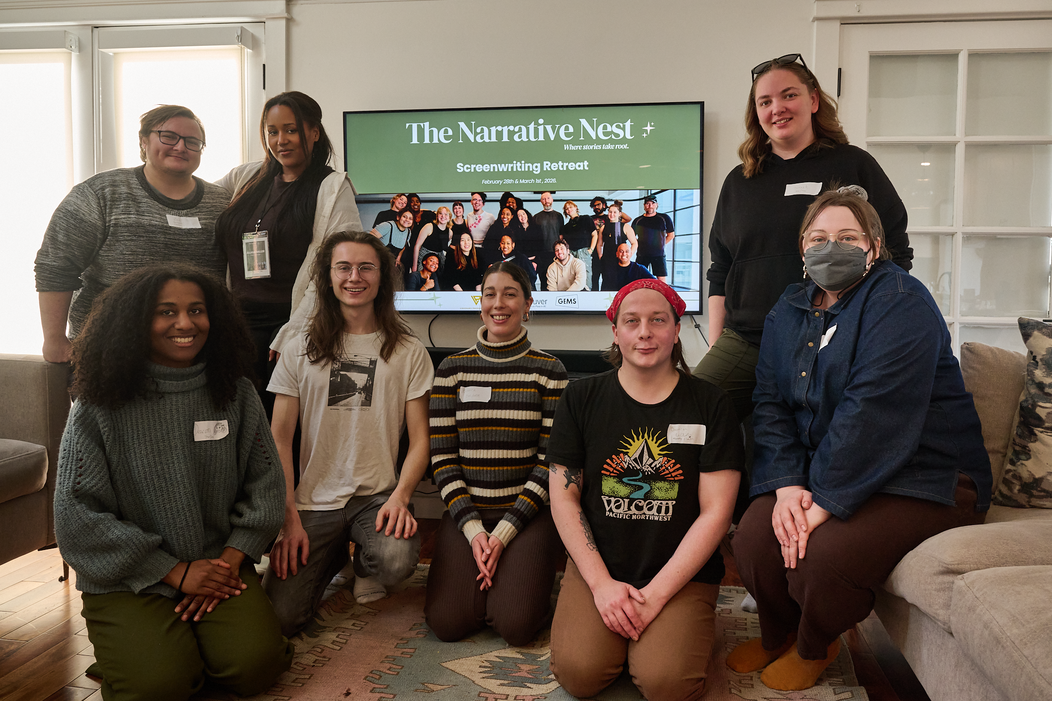 Group of eight diverse women and men posing in front of a large screen at a screenwriting retreat titled 'The Narrative Nest'.