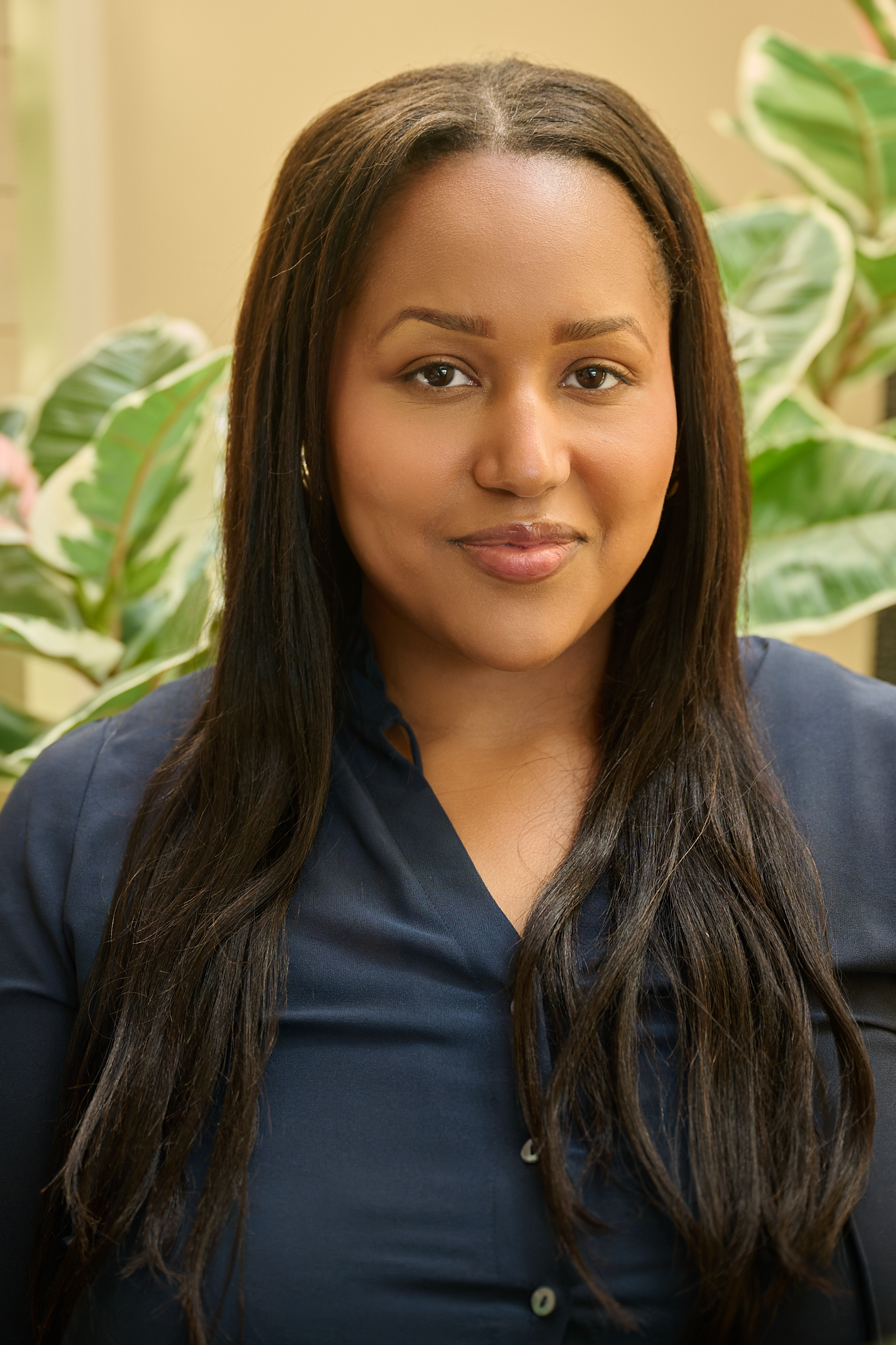 A woman with long dark hair and medium skin tone, wearing a navy blue shirt, standing indoors with large green leafy plants in the background.