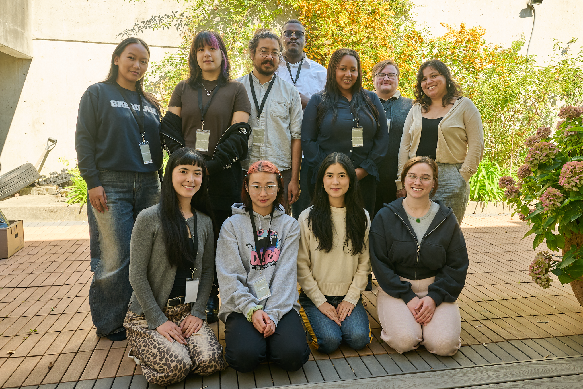 group of diverse young adults posing outdoors on a sunny day, standing and sitting on a wooden deck with green foliage and flowers in the background.