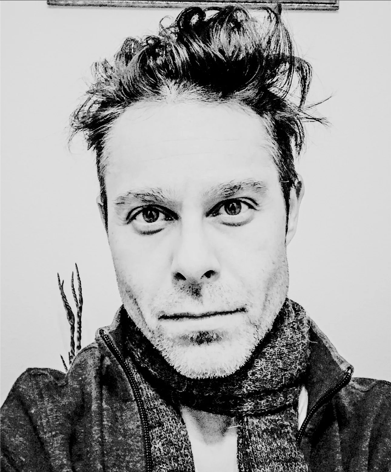 A black and white photo of a man with tousled hair, wearing a jacket and a scarf, looking directly at the camera. Jorge Heredia