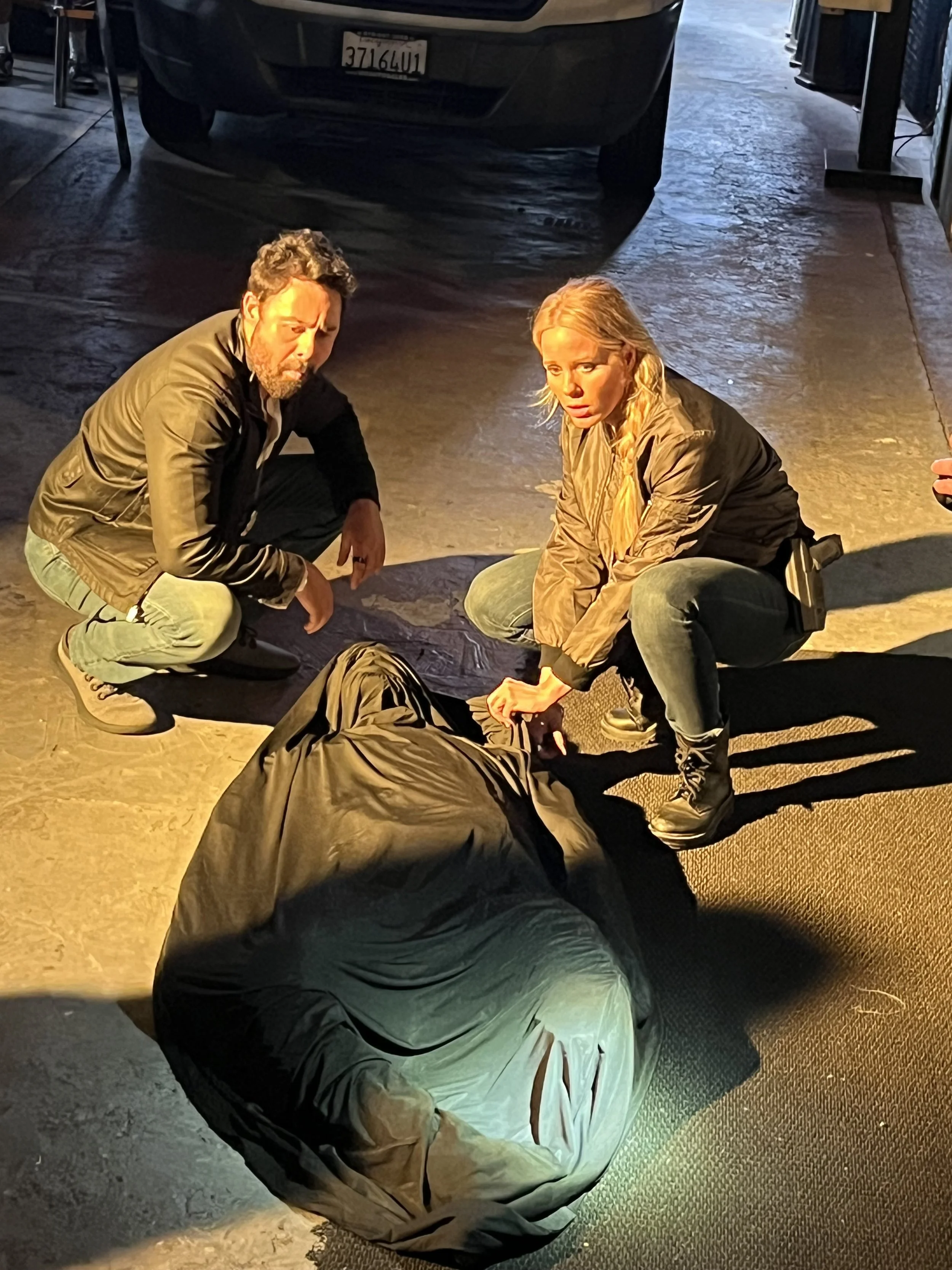 Two people, a man and a woman, squatting on the ground next to a large black bag, in a parking garage with a car overhead. Police crime scene. Jorge Heredia and LeeAnne Bauer, film Dark Mode.