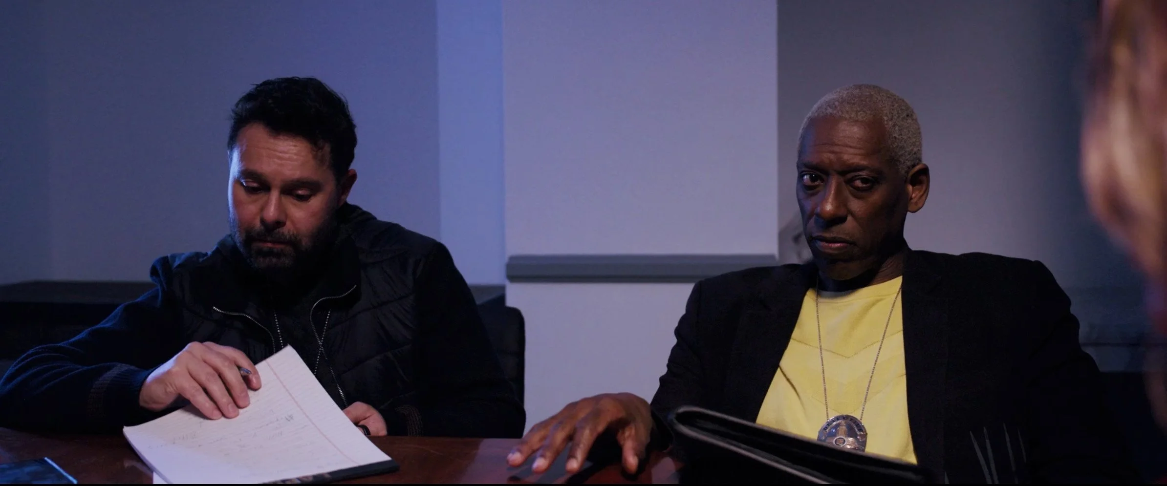 Two men sitting at a table in a dimly lit room, one with dark hair and beard looking at papers, the other with short gray hair looking serious, with a blurred person on the right side. Orlando Jones and Jorge Heredia as LAPD Detectives.