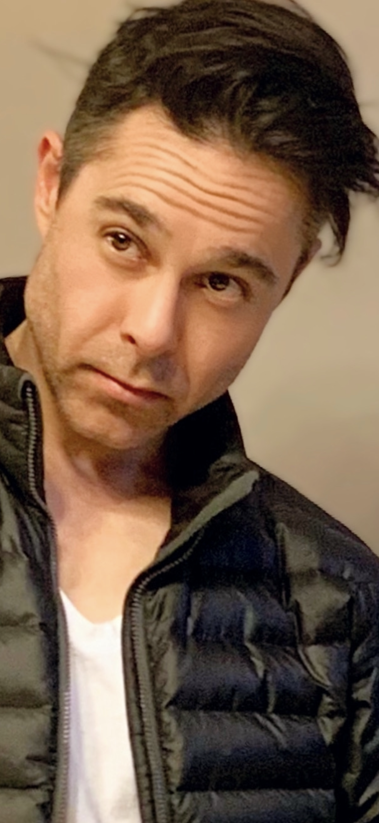 A young man with dark hair, lying on a bed, wearing a black puffy jacket and a white shirt, looking at the camera. Jorge Heredia