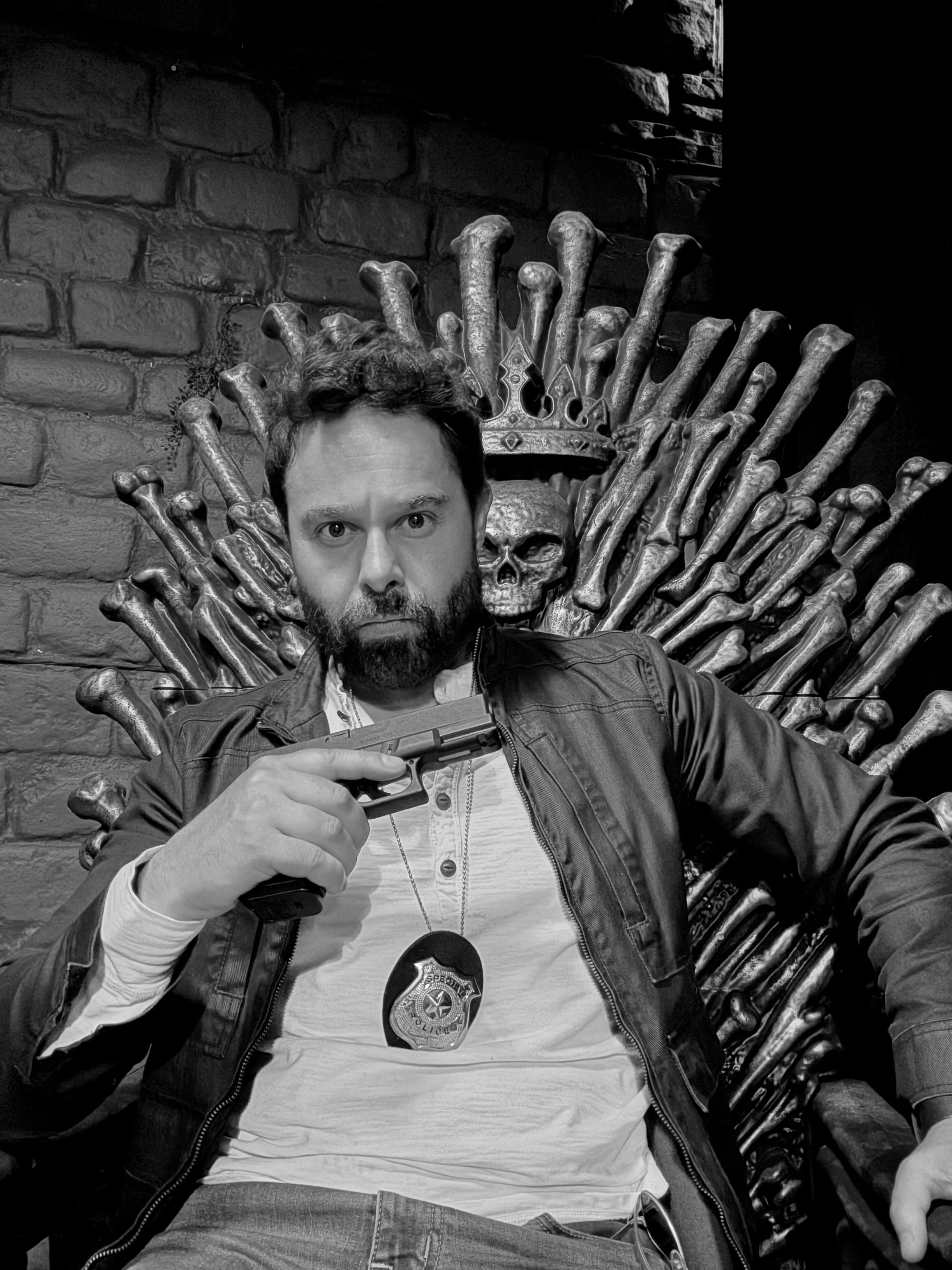 A man with a beard and hair, dressed in a leather jacket and white shirt, sitting on a throne made of bones and skulls, holding a gun, with a police badge hanging around his neck, against a brick wall.  Jorge Heredia