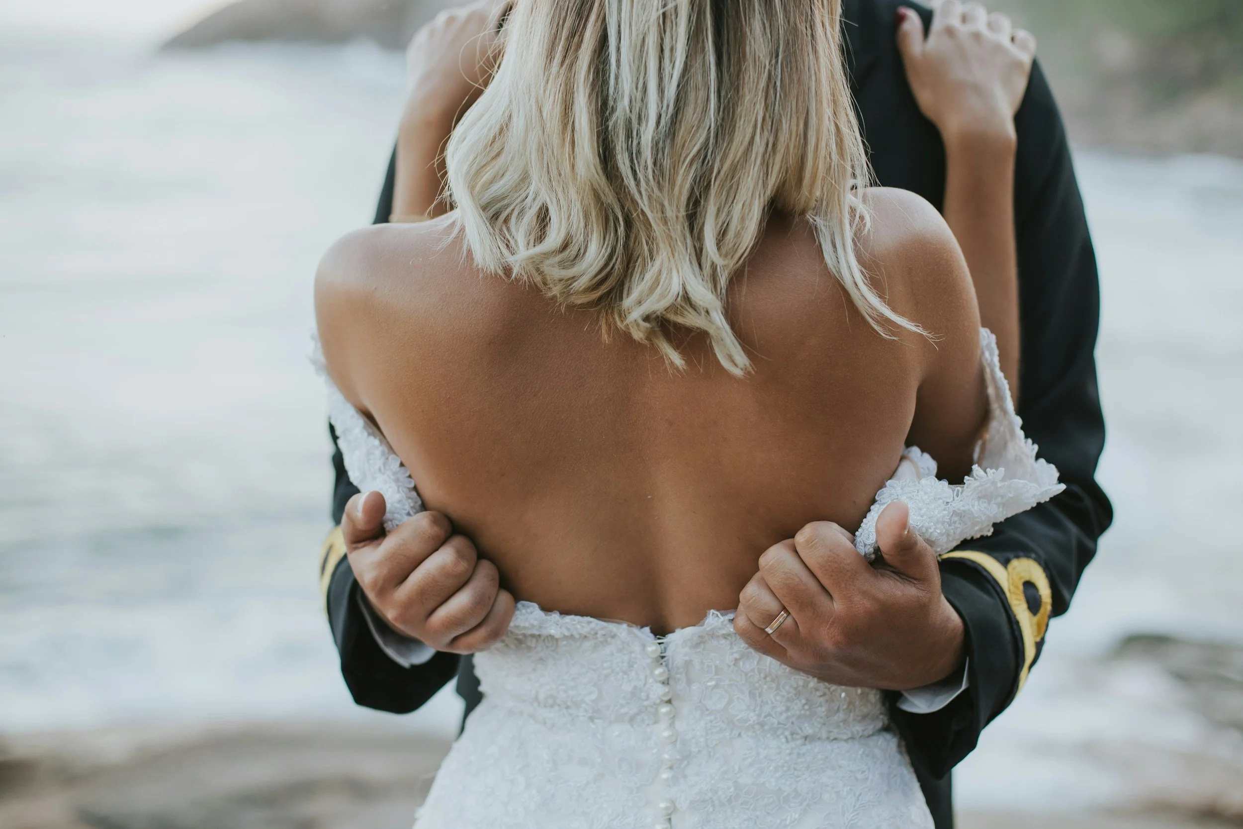 A woman in a white wedding dress being embraced by a man in a black suit. They are standing outdoors near a body of water.