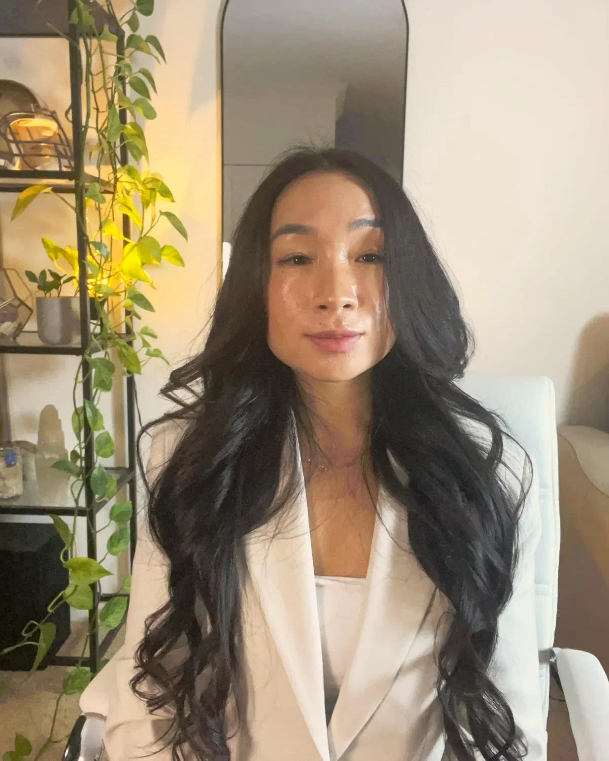 A woman with long, wavy black hair sitting in a white chair in an indoor setting, with a plant and shelves in the background.