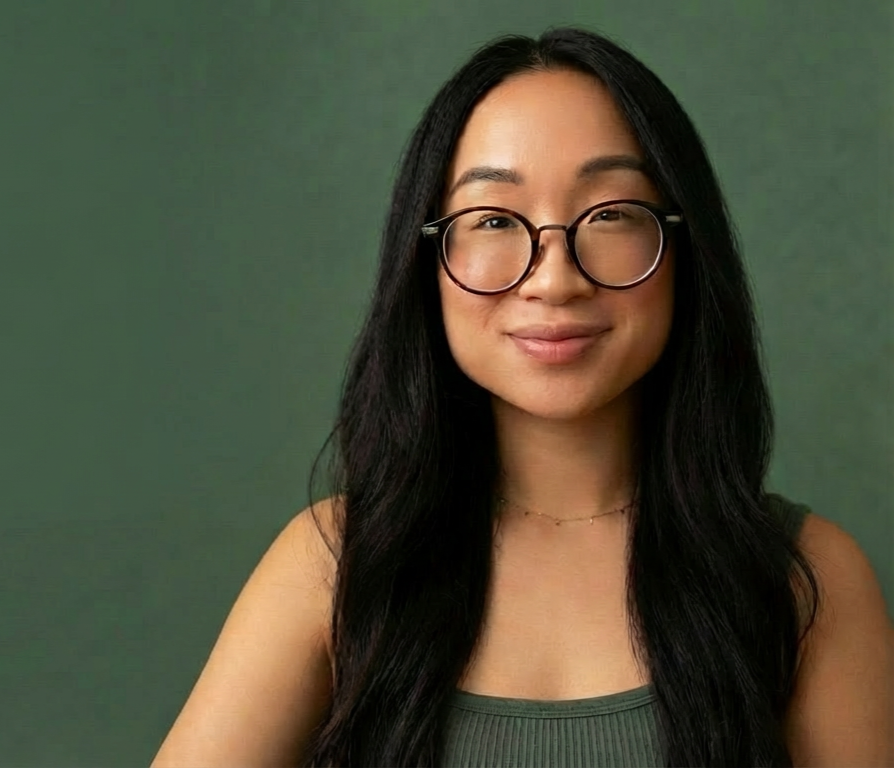 A woman with long black hair, wearing glasses and a green tank top, smiling against a green background.