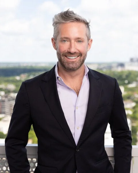 A man in a black suit and light purple shirt standing outdoors with a cityscape and blue sky in the background.