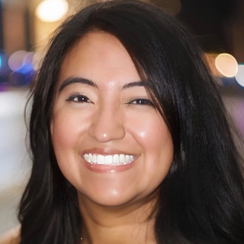 A woman with long black hair smiling warmly at the camera against a blurred background with lights.
