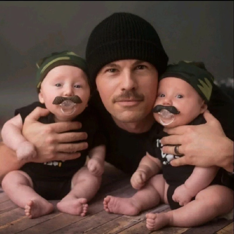 Man lying on the floor with two babies, each wearing a black beanie and black clothing, with fake mustache and pacifier, in a close-up portrait.