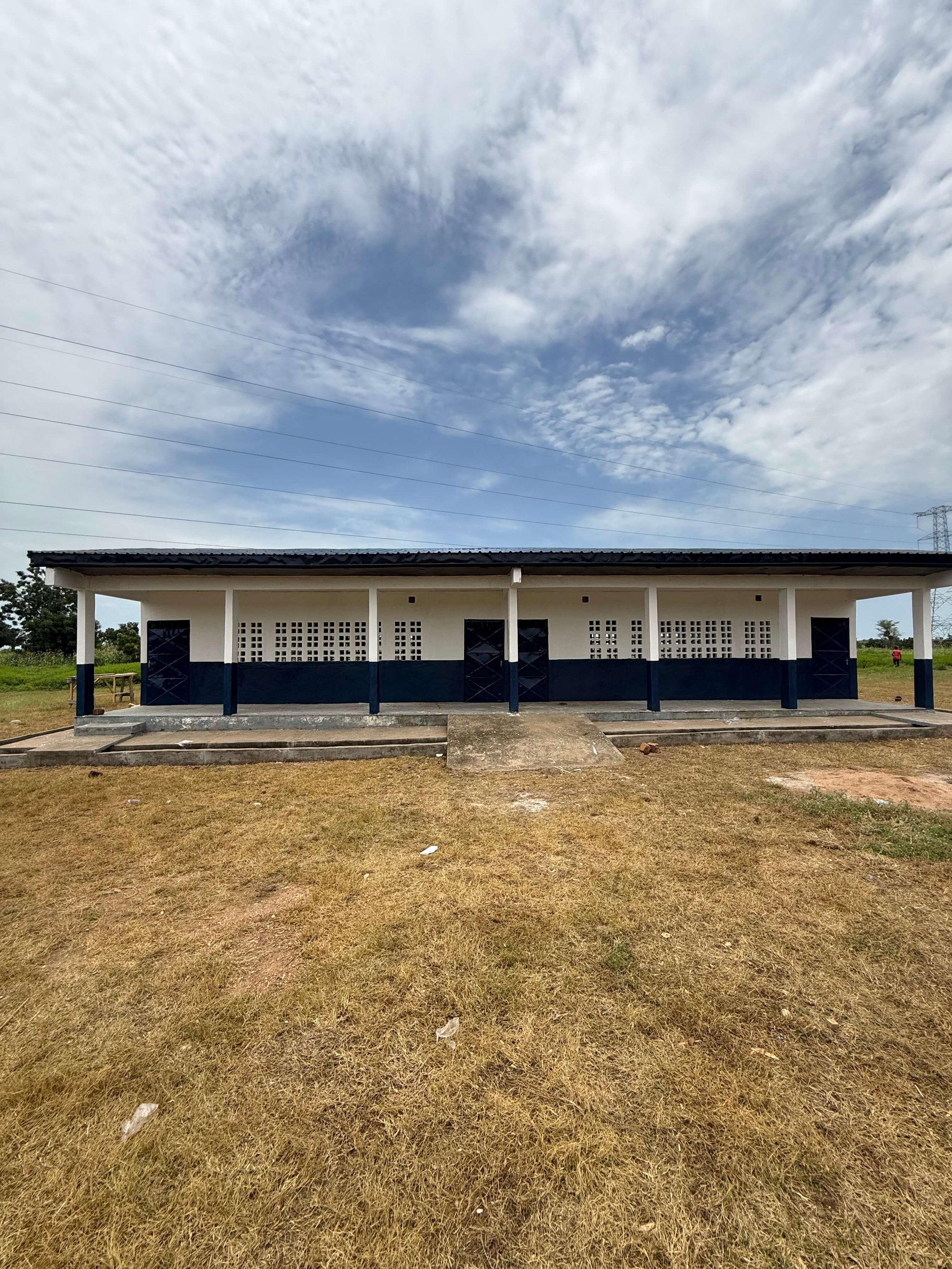 Long blue-and-white open-air building with concrete walkway, dry grass, cloudy sky, and power lines