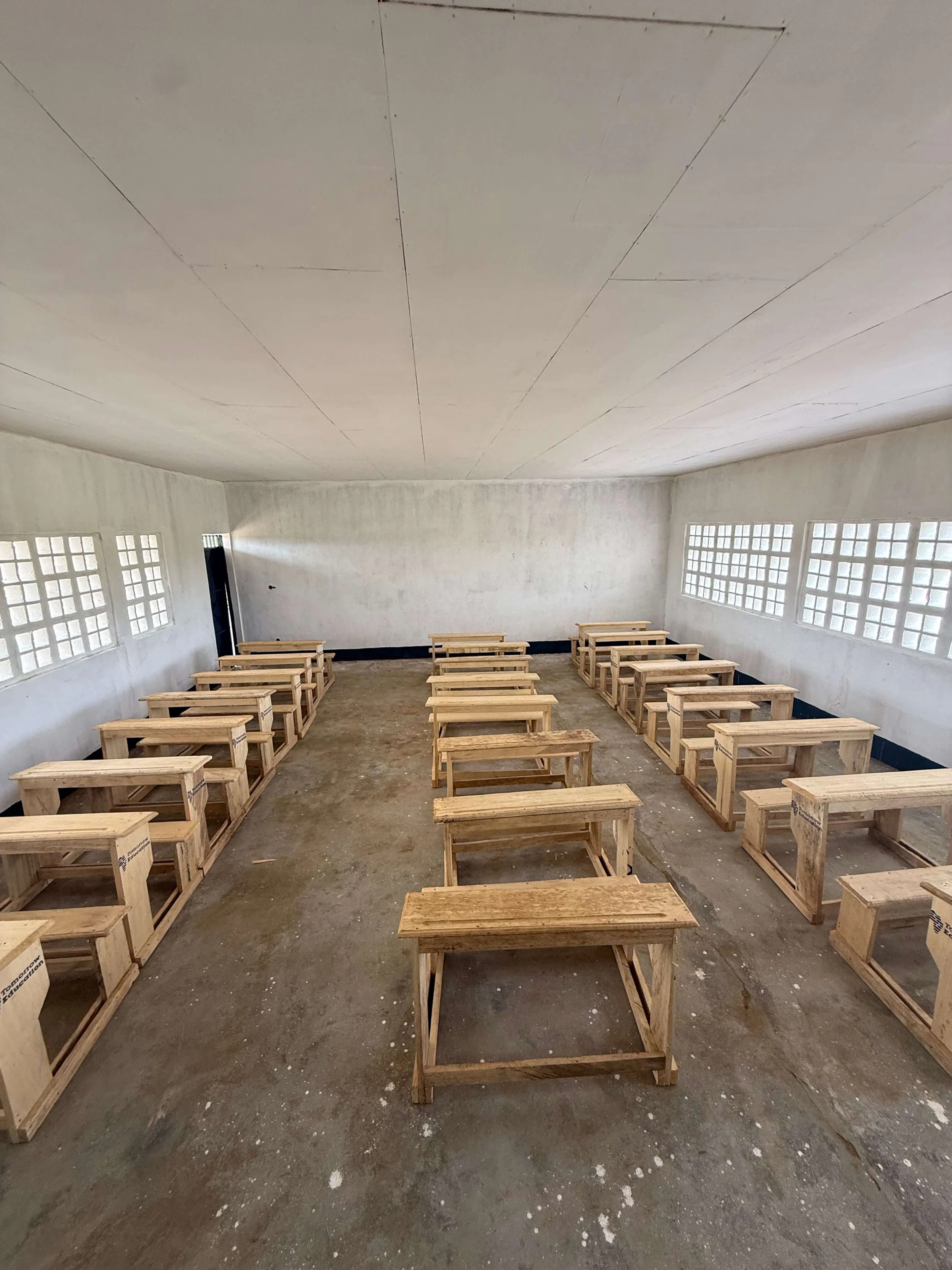 Empty classroom with rows of wooden student desks, concrete walls, and glass-block windows