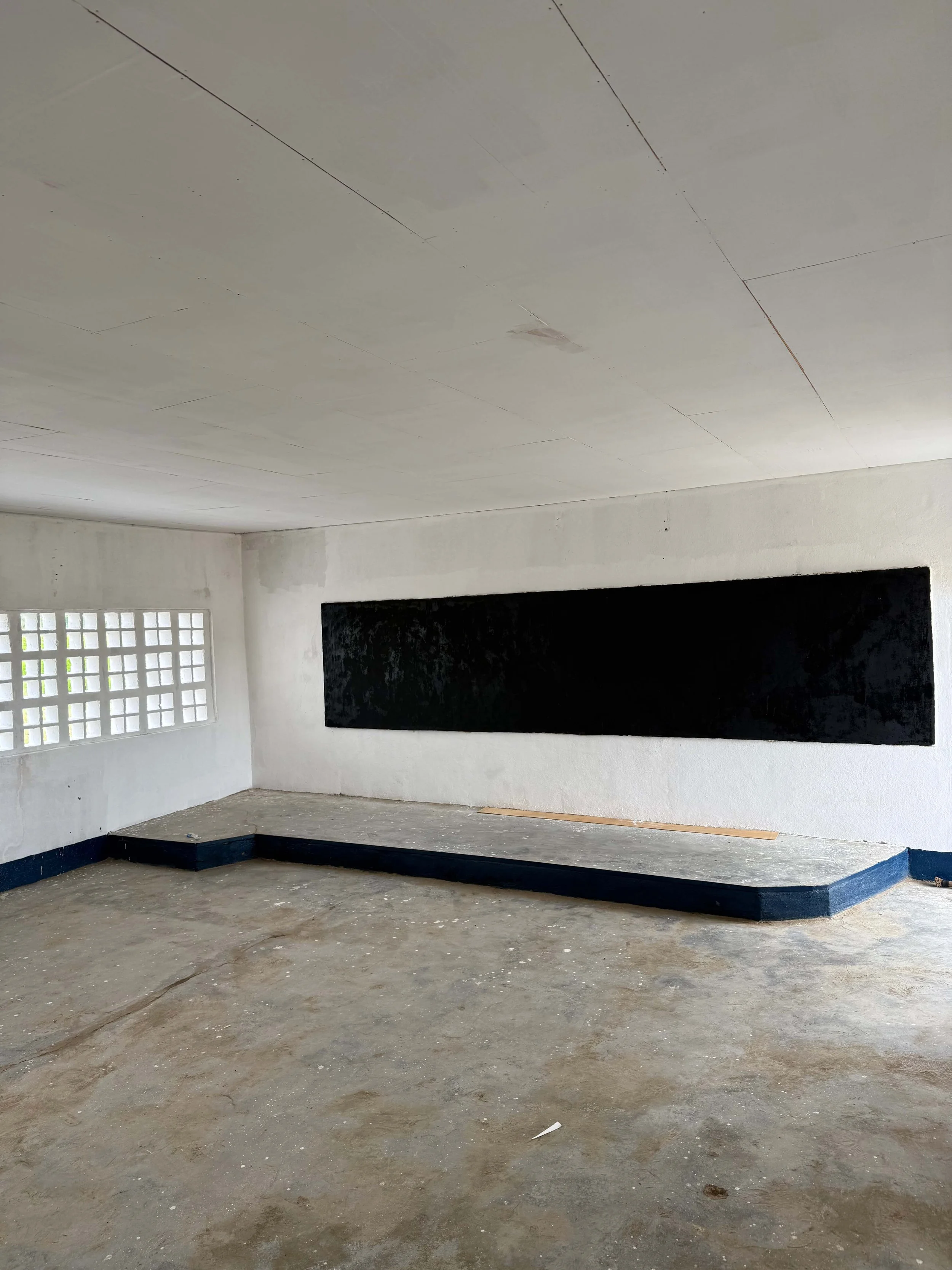 Empty unfinished interior with a raised concrete stage, black-painted wall opening, glass-block window, and worn concrete floor