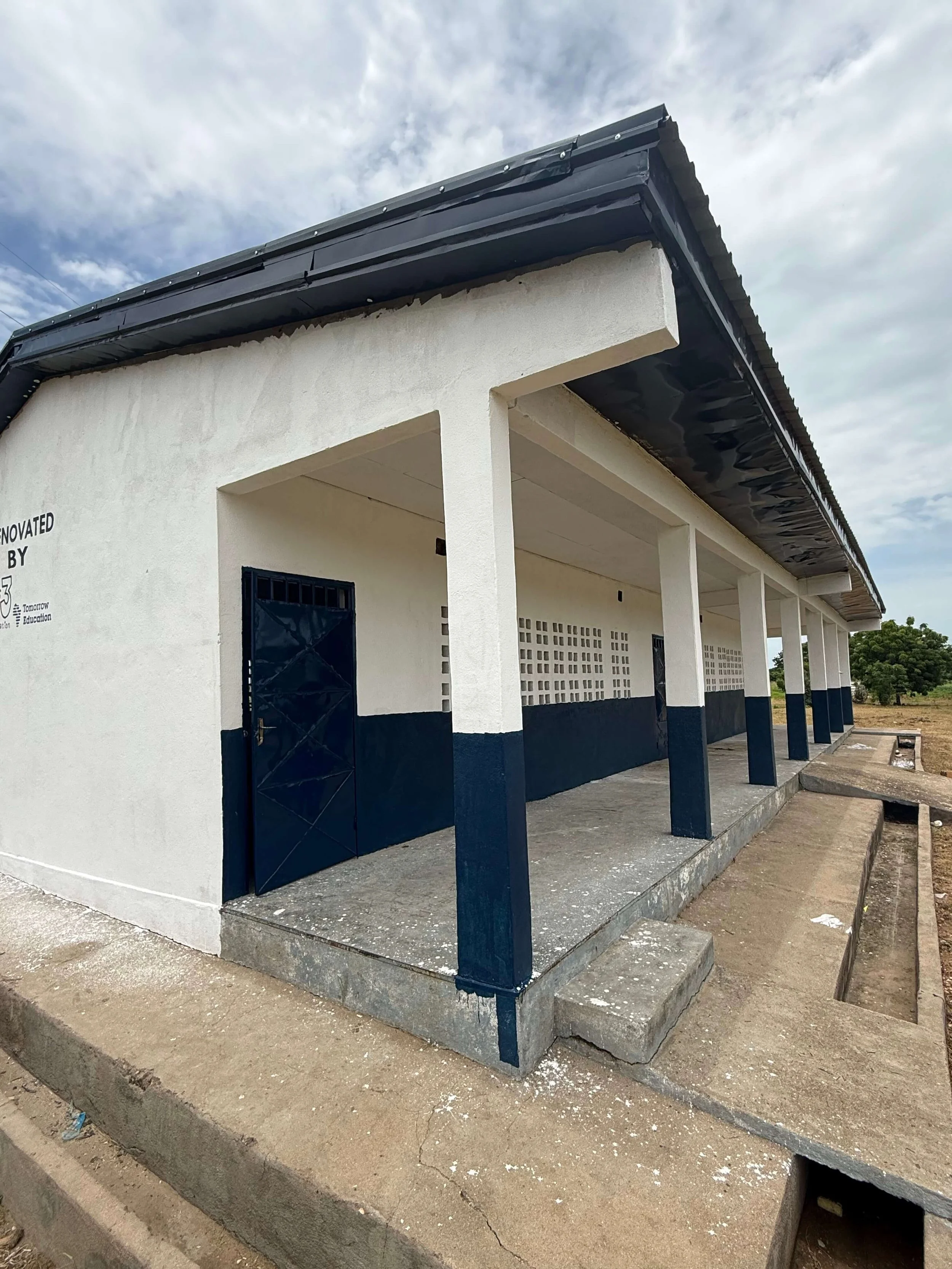 White-and-navy community school building with covered walkway, black doors, and damaged roof under a cloudy sky