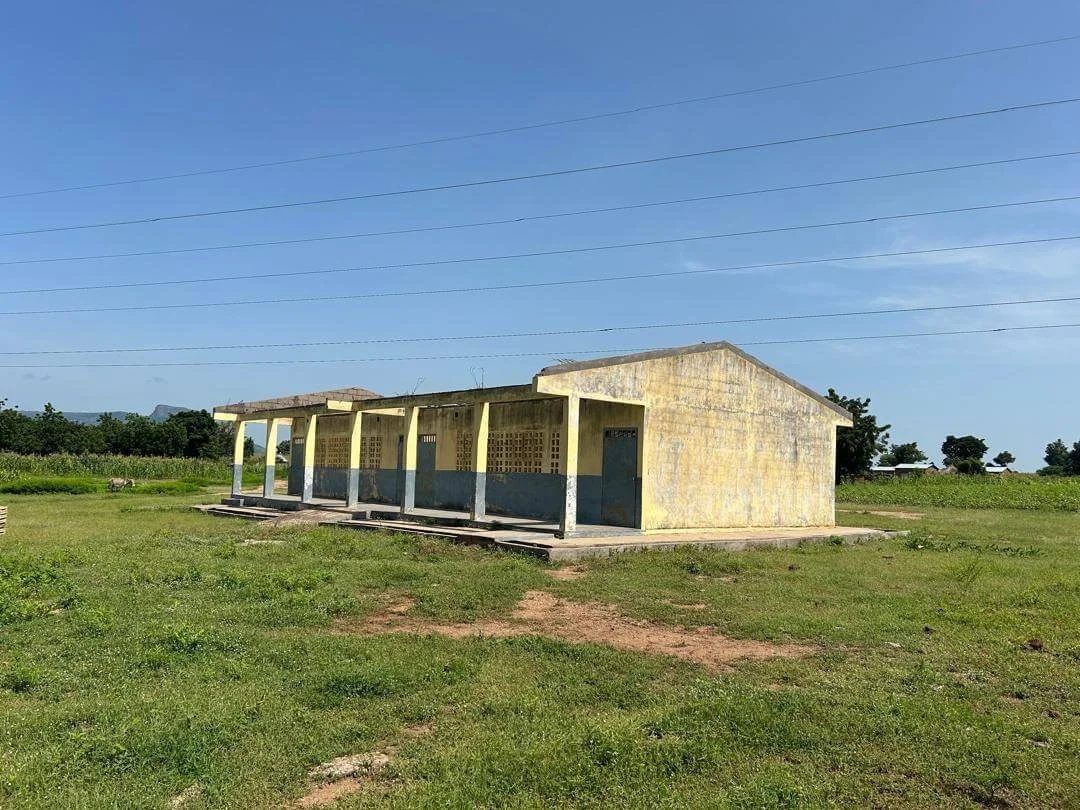 Weathered yellow concrete building with a covered porch in a grassy rural field under a clear blue sky