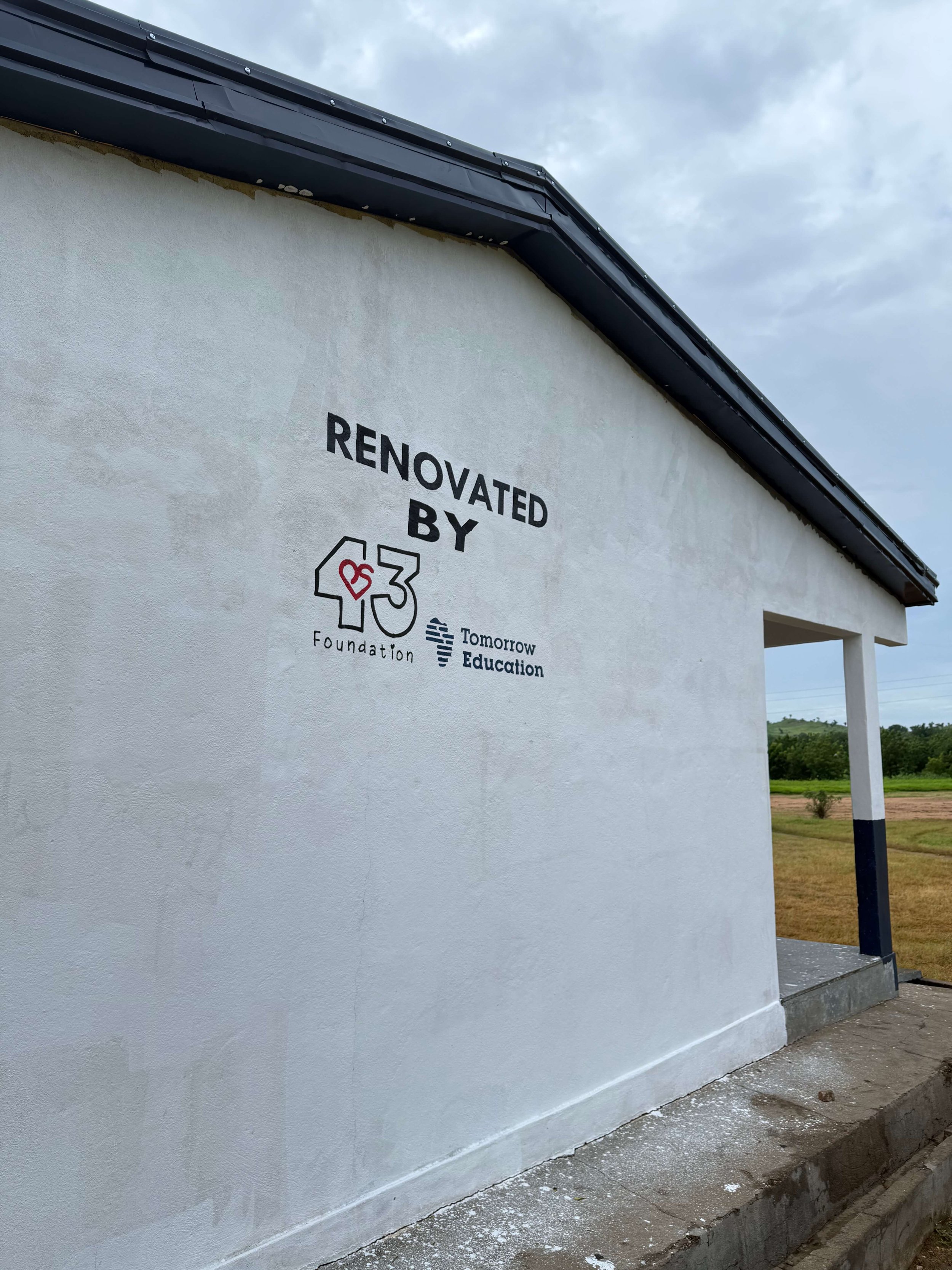 White school building with black roof trim displaying a "Renovated by PS43 Foundation" and "Tomorrow Education" logo beside a rural field.
