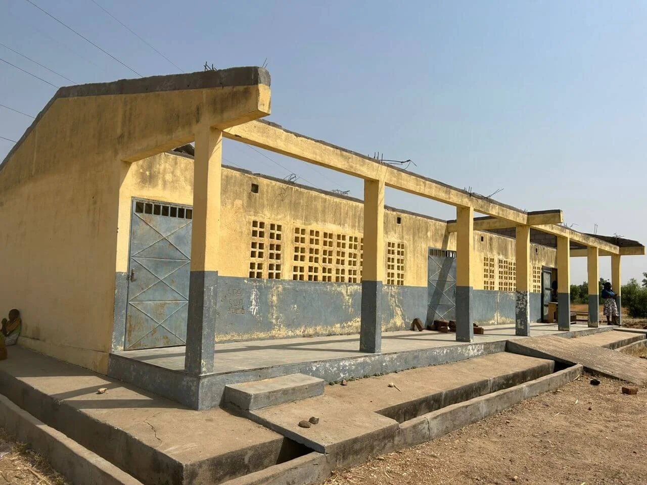 Weathered yellow-and-gray concrete building with covered walkway, metal doors, ventilation blocks, and open drainage channels