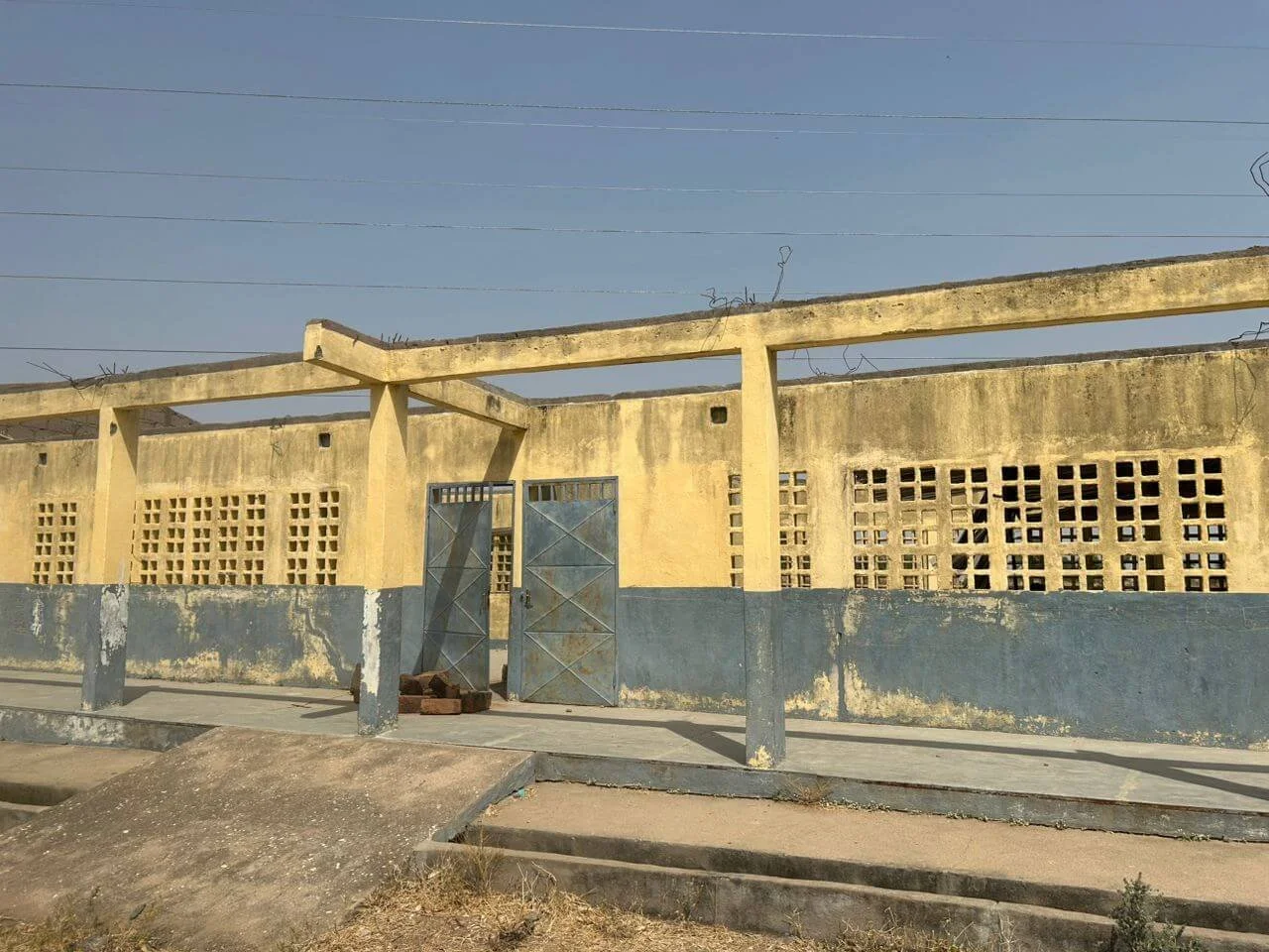 Weathered yellow-and-blue concrete building with metal gates, ventilation blocks, and exposed roof beams beside a street