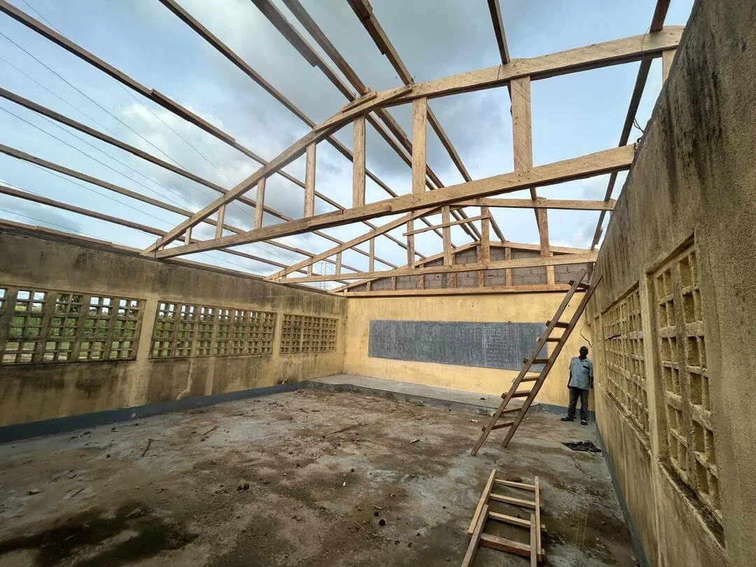 Unfinished schoolroom with exposed wooden roof beams, concrete walls, a chalkboard, glass-block windows, and a ladder.