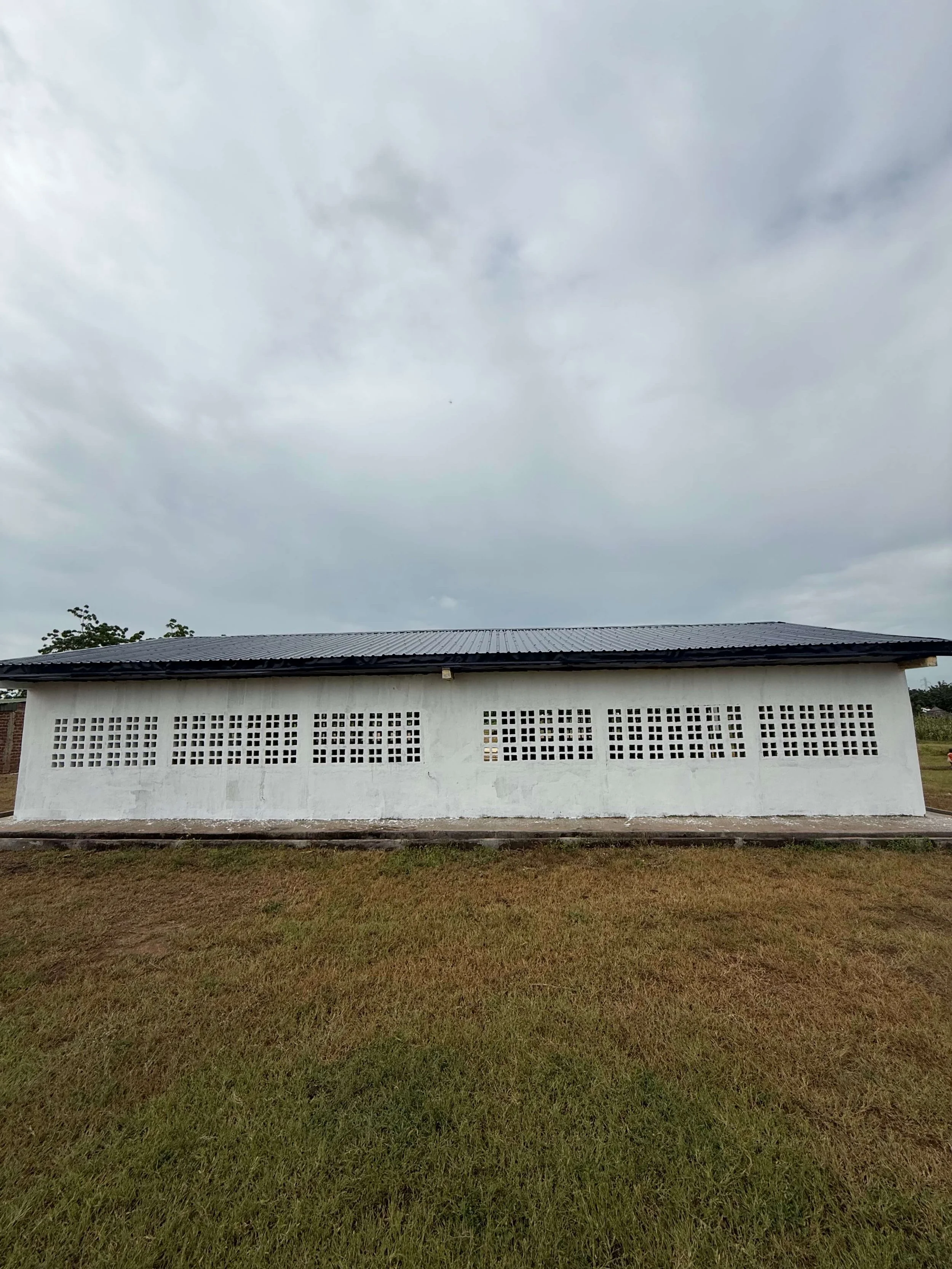 White concrete building with patterned ventilation blocks and corrugated metal roof beside a grassy field under an overcast sky