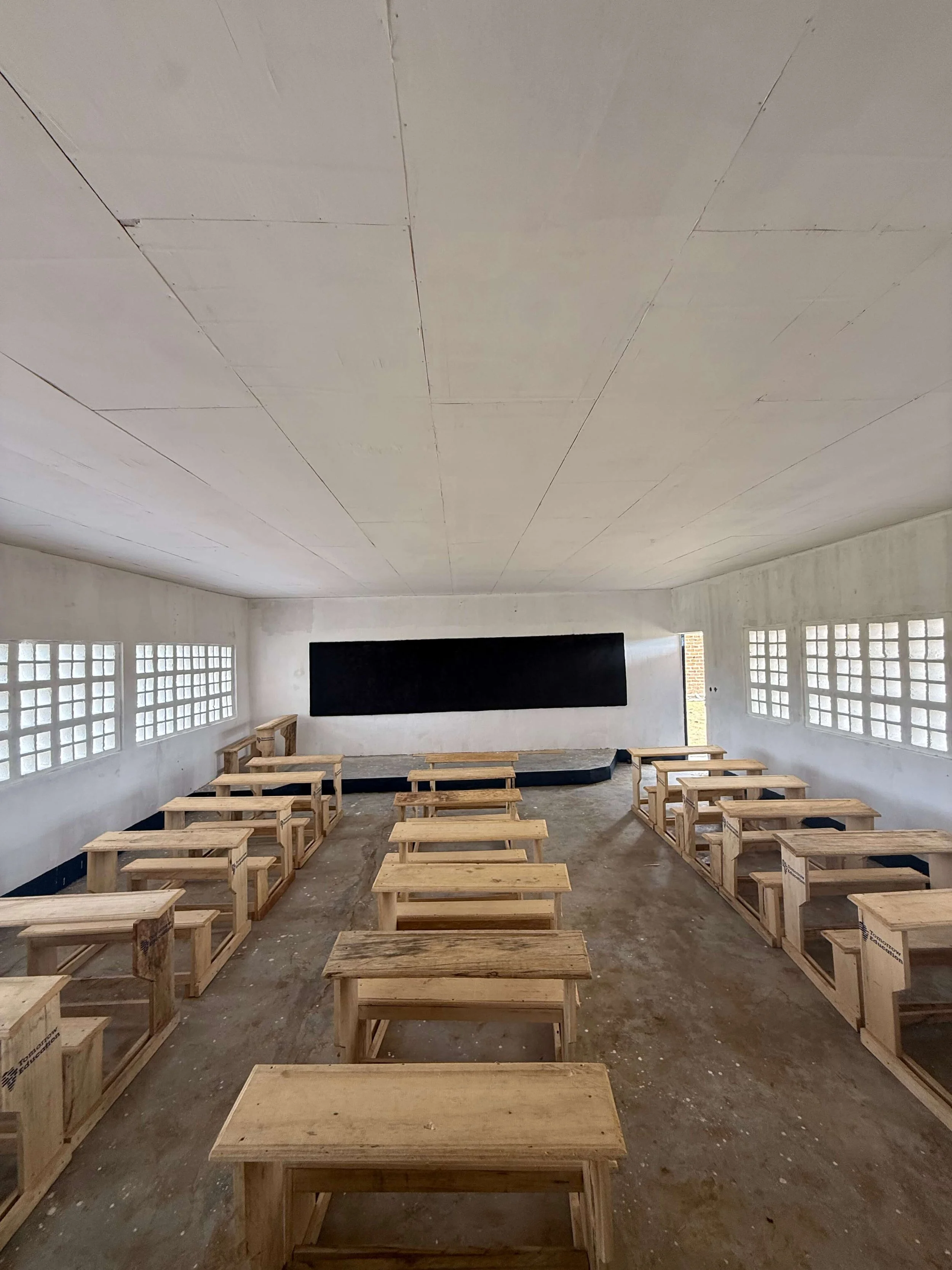 Empty classroom with rows of unfinished wooden desks, glass-block windows, and a blackboard